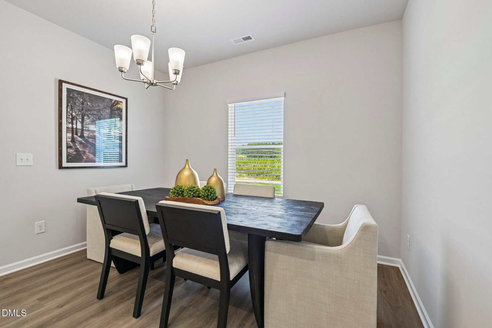 Modern dining room with a black table, three black chairs, and a cream upholstered bench. A window lets in light, and a chandelier hangs above. Art with trees and gold vases decorate the space.