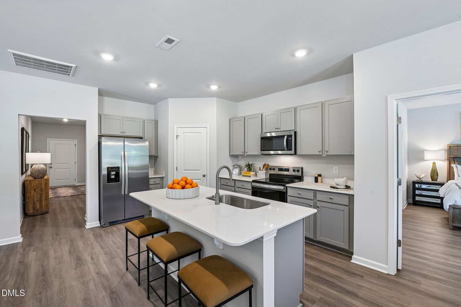 Modern kitchen with gray cabinets, stainless steel appliances, a center island with three stools, and a bowl of oranges. An open doorway leads to a bedroom, and another to a hallway with a table and lamp.