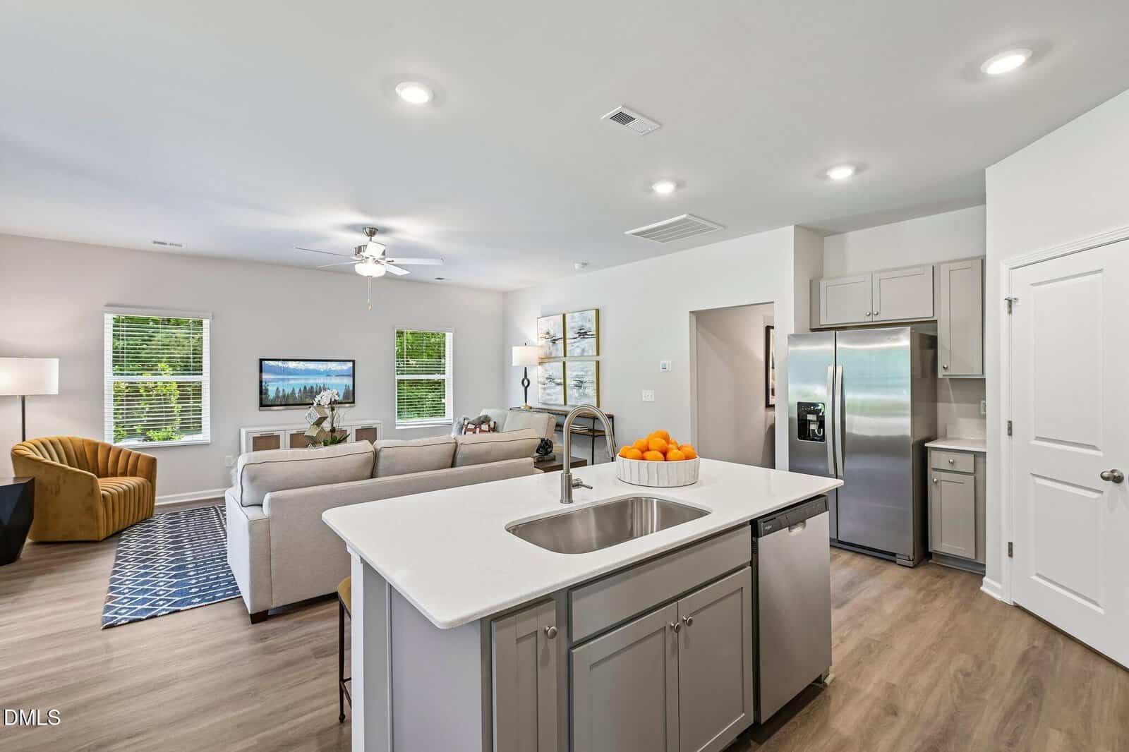 Modern open-concept kitchen and living room with a white island, stainless steel appliances, light wood floors, neutral-colored furniture, and large windows letting in natural light. Bowl of oranges sits on the island.