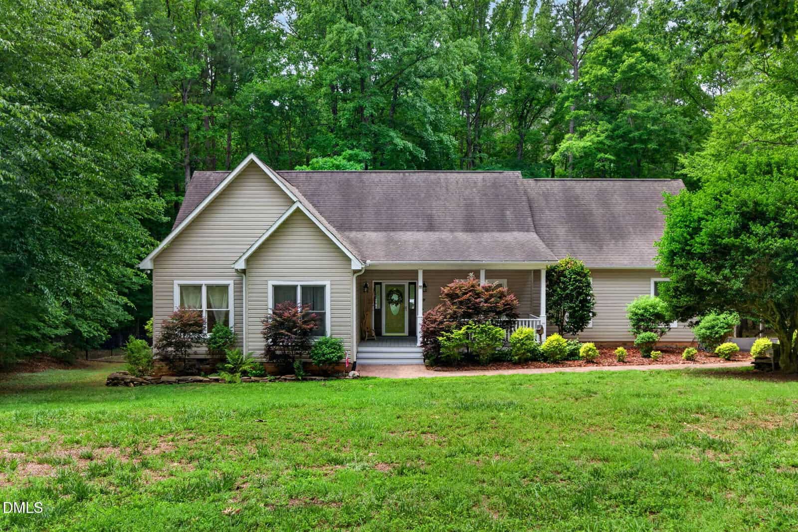 A single-story beige house on 475 Dreamcatcher Trail in Youngsville sits surrounded by lush green trees and shrubs, featuring a front porch and a well-maintained lawn.