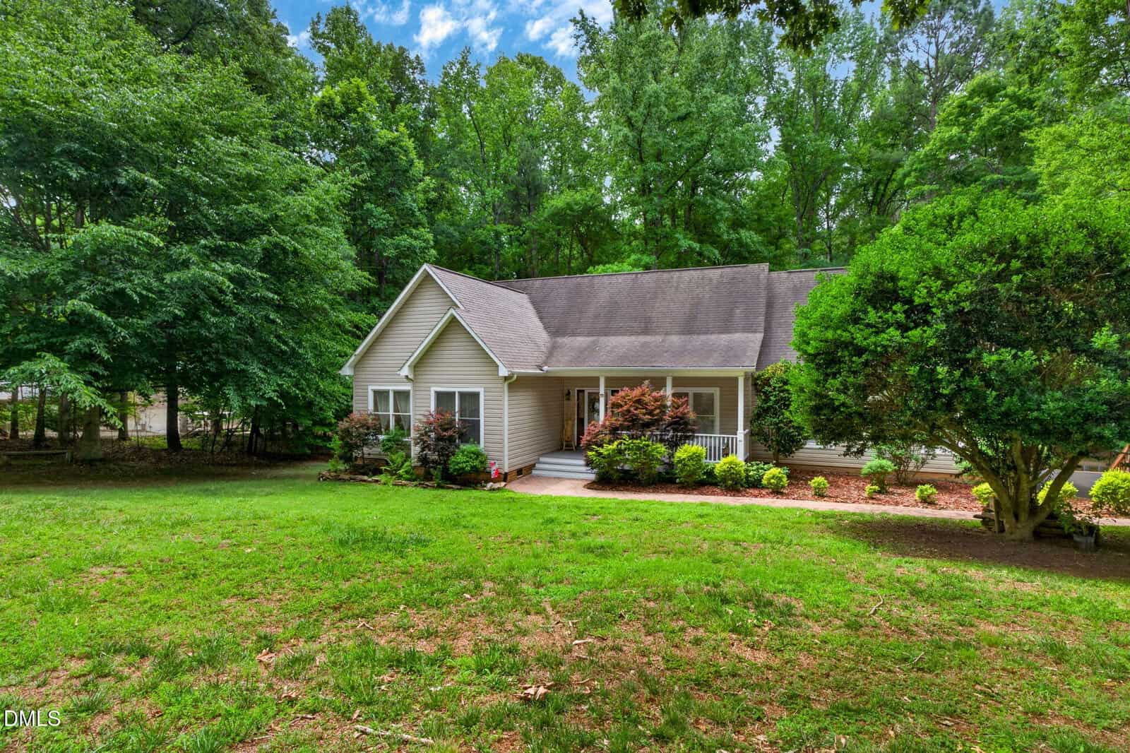 A single-story house with a gable roof surrounded by lush green trees and a well-maintained lawn, featuring a small front porch and neatly trimmed bushes and shrubs in the garden.