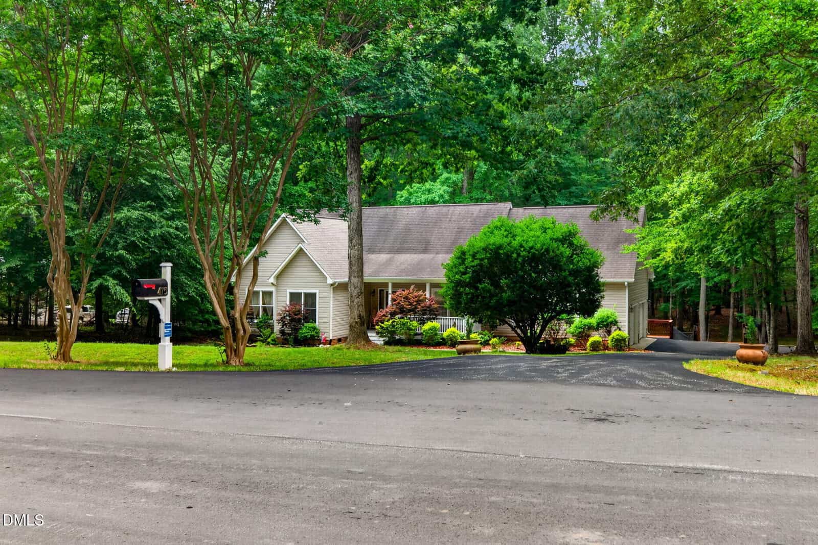 A single-story house with light-colored siding, surrounded by green trees and shrubs, sits behind a paved driveway and mailbox on a quiet asphalt road. Large potted plants decorate the front yard.