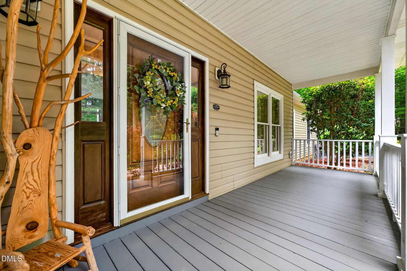 Covered front porch with light tan siding, gray floorboards, a wooden door with a glass storm door decorated with a floral wreath, a wooden chair, white railing, and a view of greenery.