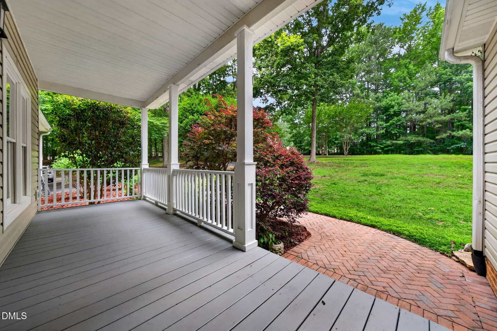 Covered porch with gray wooden floor, white railing, and columns overlooks a brick walkway, lush green lawn, and trees in the background on a bright day.