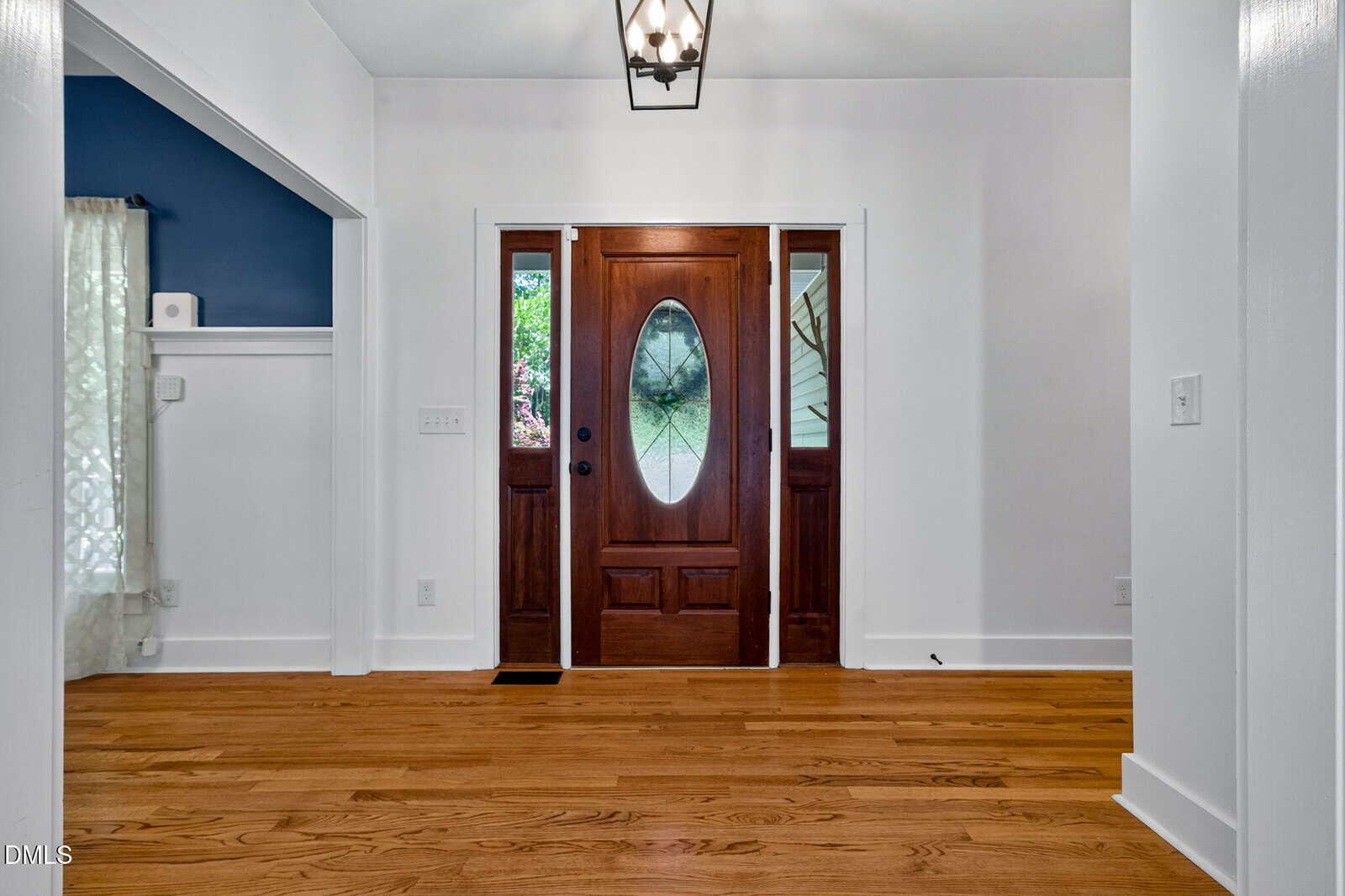 A wooden front door with two glass panels stands in a bright entryway with white walls and hardwood floors. A hanging light fixture is overhead, and a glimpse of a blue room is visible to the left.