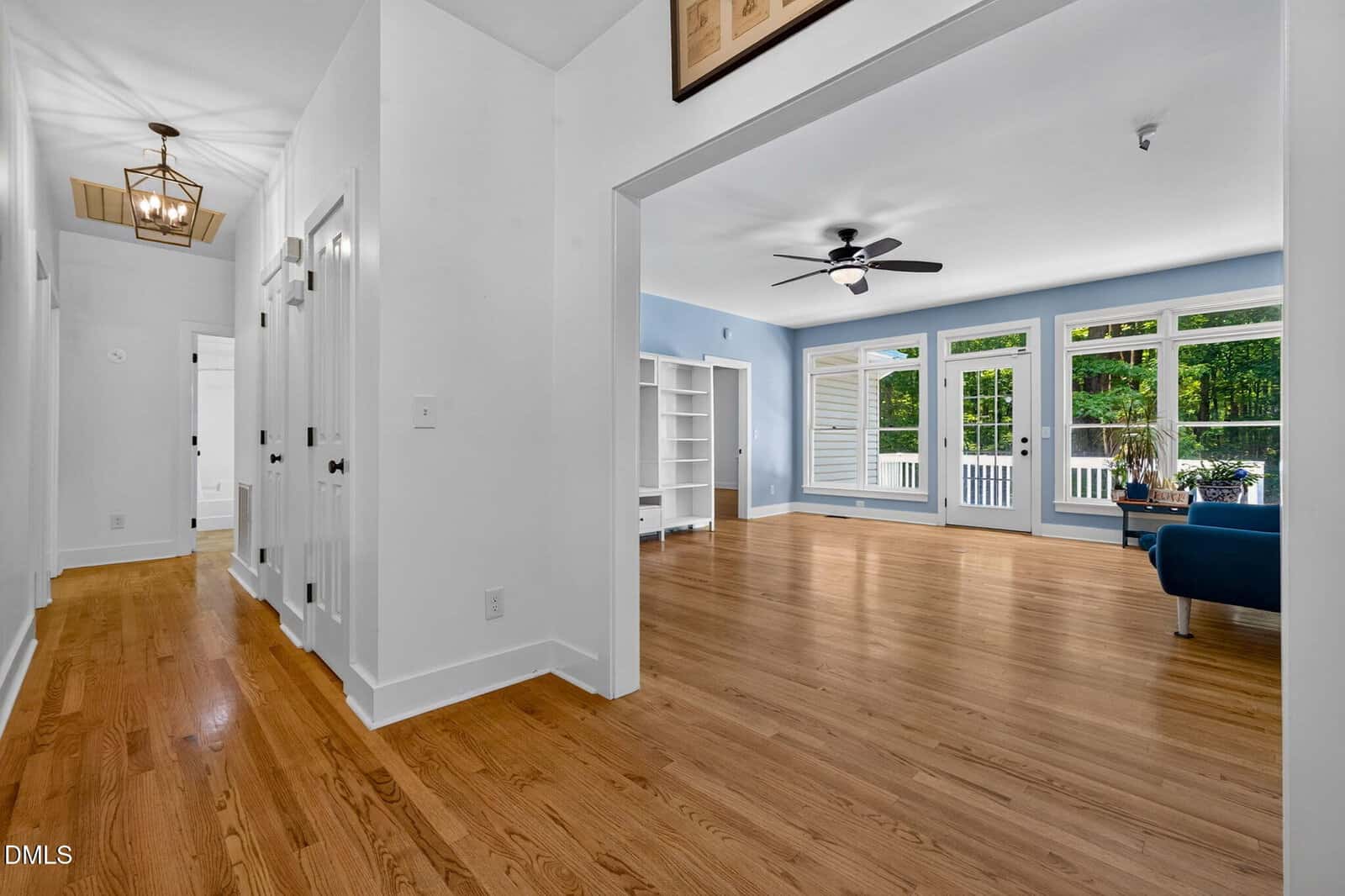 Bright hallway with wooden floors leads to a spacious living room featuring large windows, a ceiling fan, blue walls, and minimalist furniture, including a blue sofa and white shelving.
