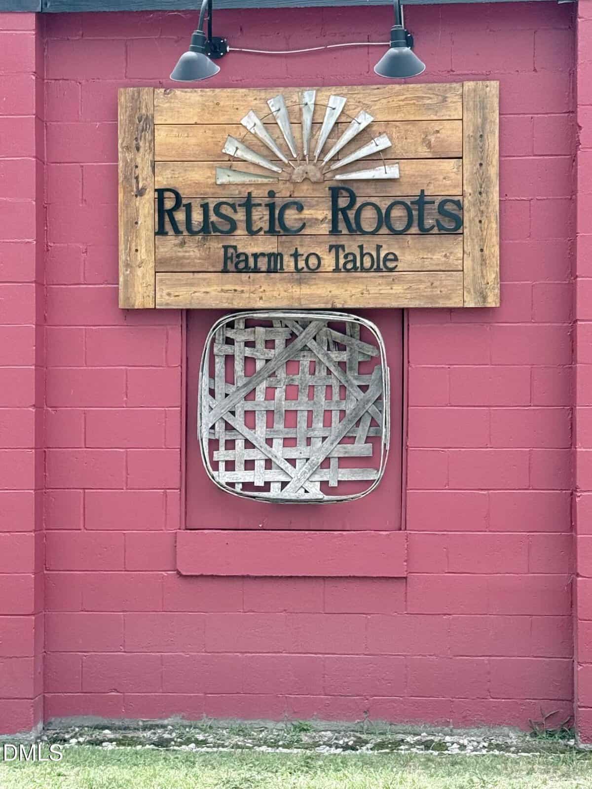 A wooden sign reading “Rustic Roots Farm to Table” is mounted on a red brick wall above a metal decorative piece. Two black lamps are fixed above the sign. Grass grows along the bottom edge of the wall.