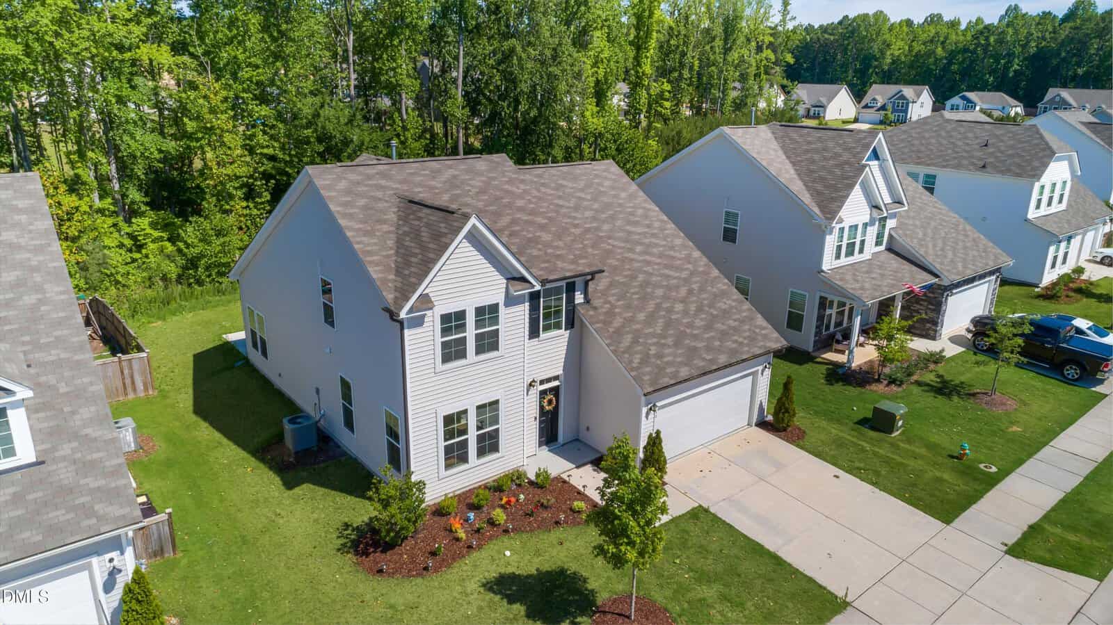 Aerial view of a two-story suburban house with gray siding, a two-car garage, manicured lawn, landscaped front yard, and neighboring houses on both sides, bordered by a wooded area in the background.