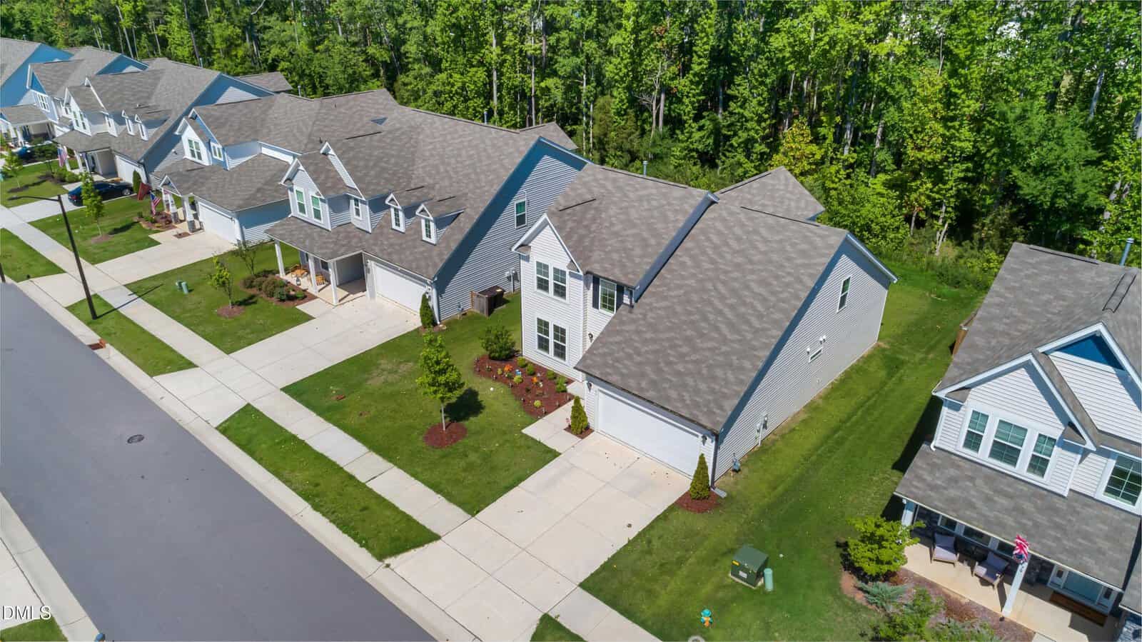 Aerial view of a suburban neighborhood with rows of modern, two-story houses, manicured lawns, driveways, and a tree-lined wooded area behind the homes.