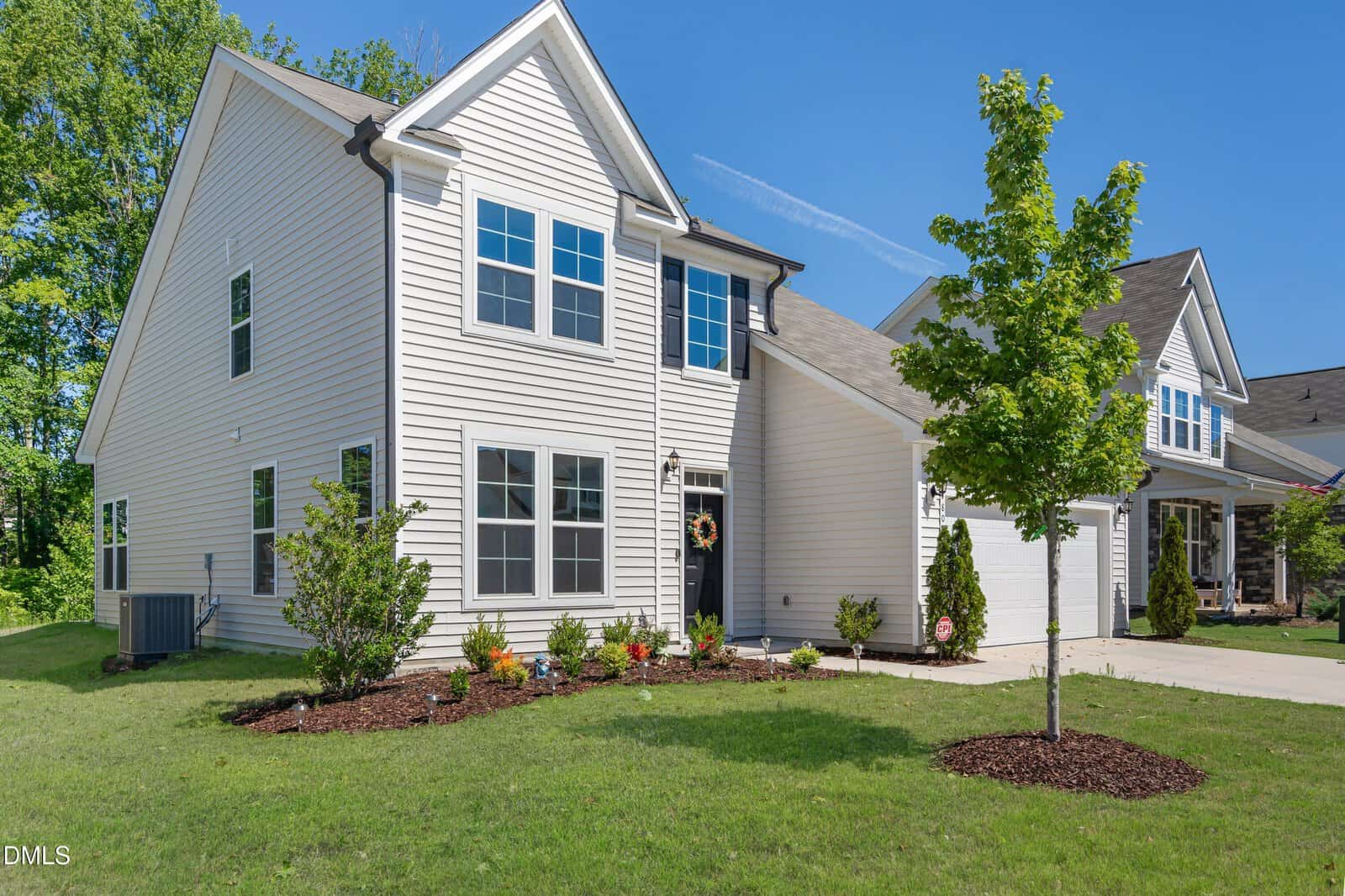 A modern two-story white house with large windows, landscaped front yard, small trees, and a driveway, set on a sunny day with a clear blue sky.