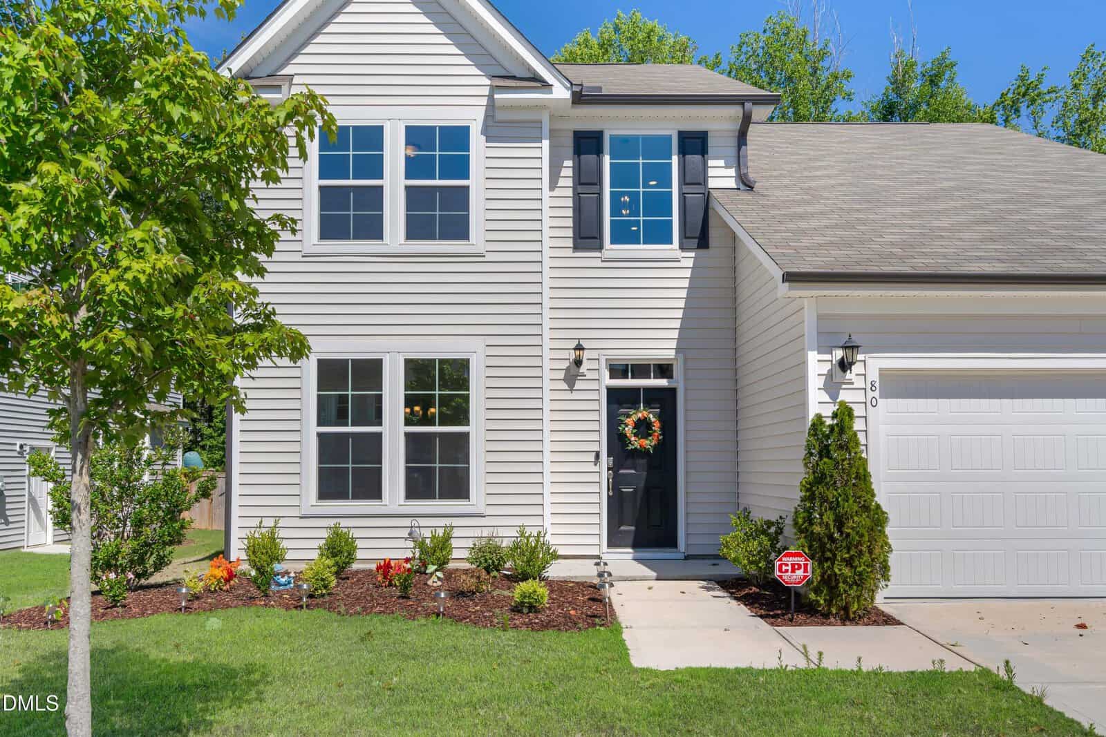 A two-story beige house with black shutters at 80 Silent Brook Trail, Franklinton, features a black front door with a wreath, an attached garage, and a landscaped front yard with shrubs and a tree on a sunny day.