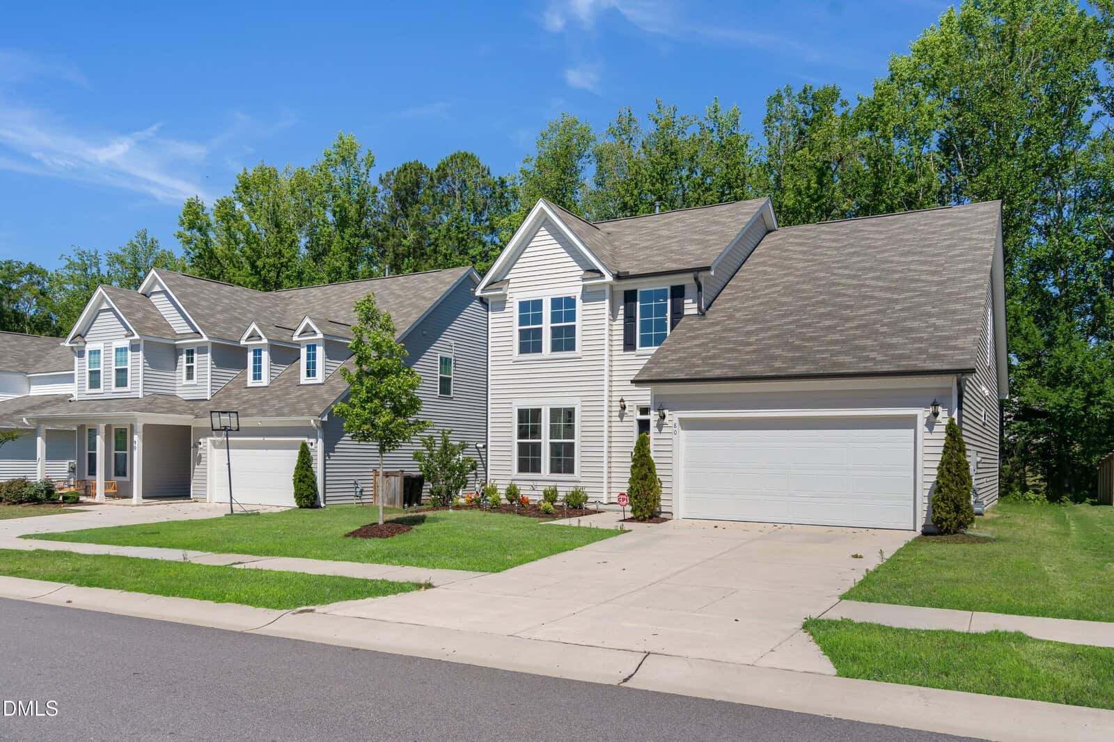 A modern, two-story suburban house with light gray siding, white trim, a two-car garage, green lawn, and young trees, set in a quiet neighborhood on a sunny day.