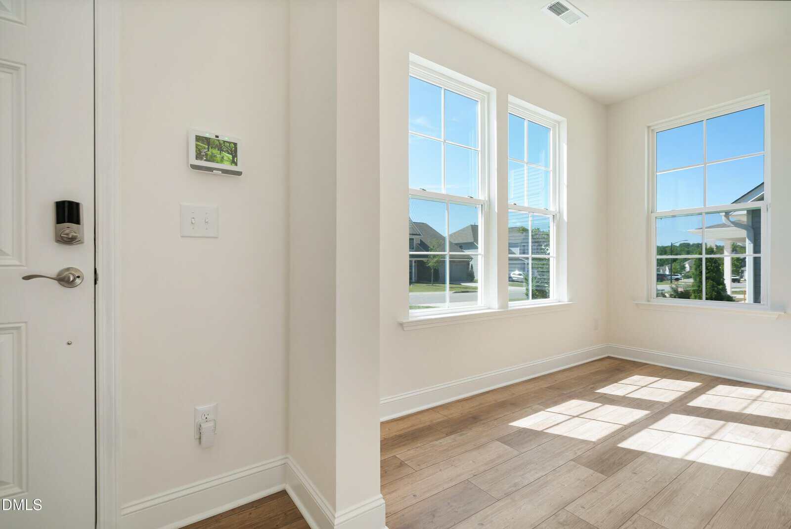 Sunlit room with light wood floors, white walls, large windows, and a white door. A thermostat and outlets are on the wall. Bright natural light fills the space.