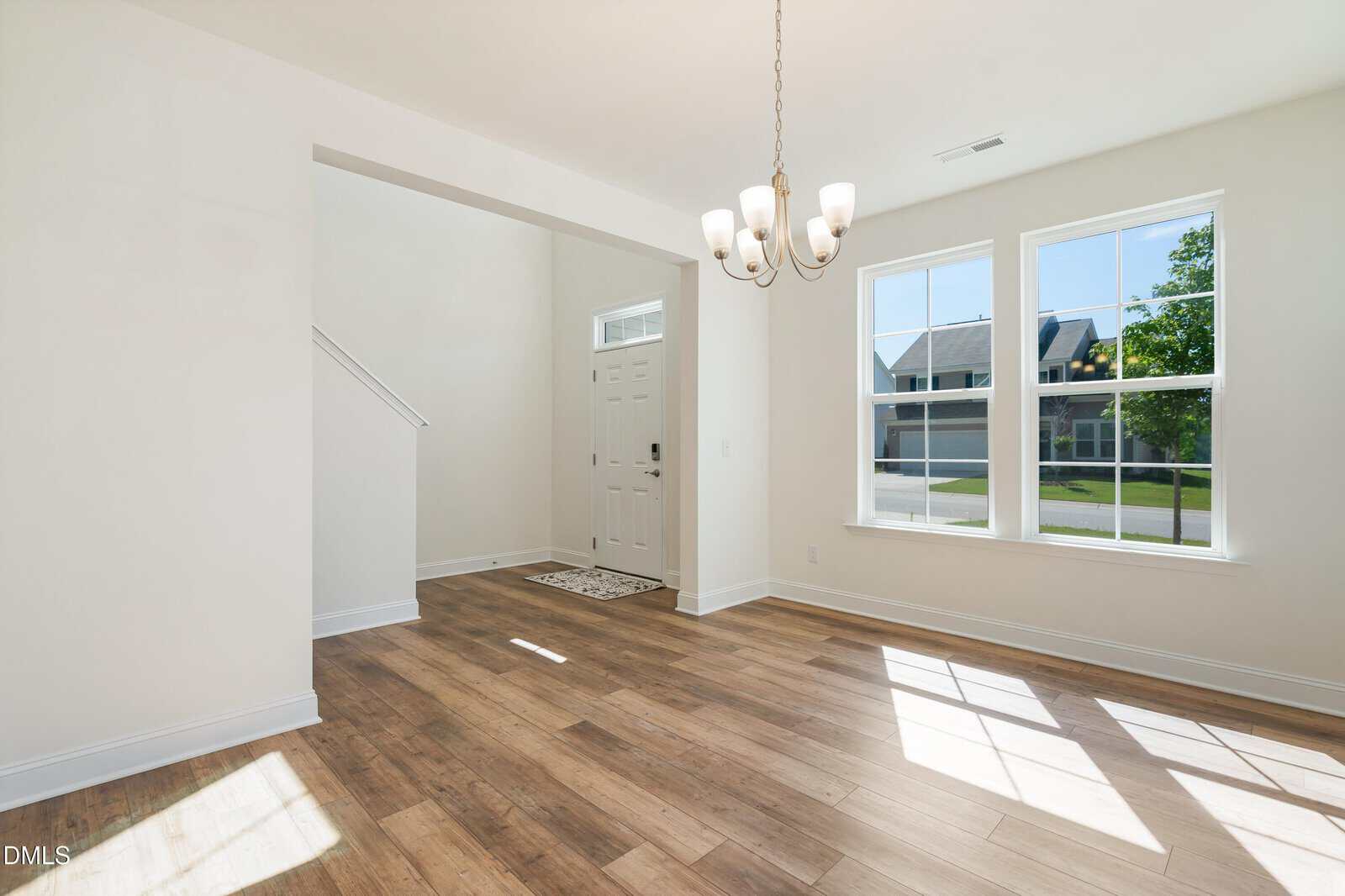 Bright, empty room with wood floors, white walls, large windows letting in sunlight, a chandelier, and a white front door visible through an open entryway.