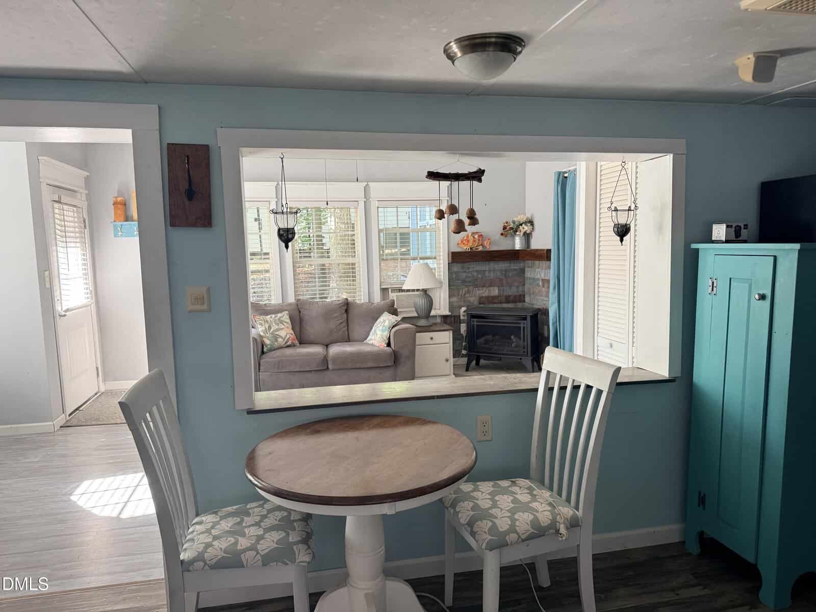 Small dining area with a round table and two chairs, set by a wide pass-through window looking into a cozy living room with a gray sofa, brick fireplace, and large windows letting in sunlight.