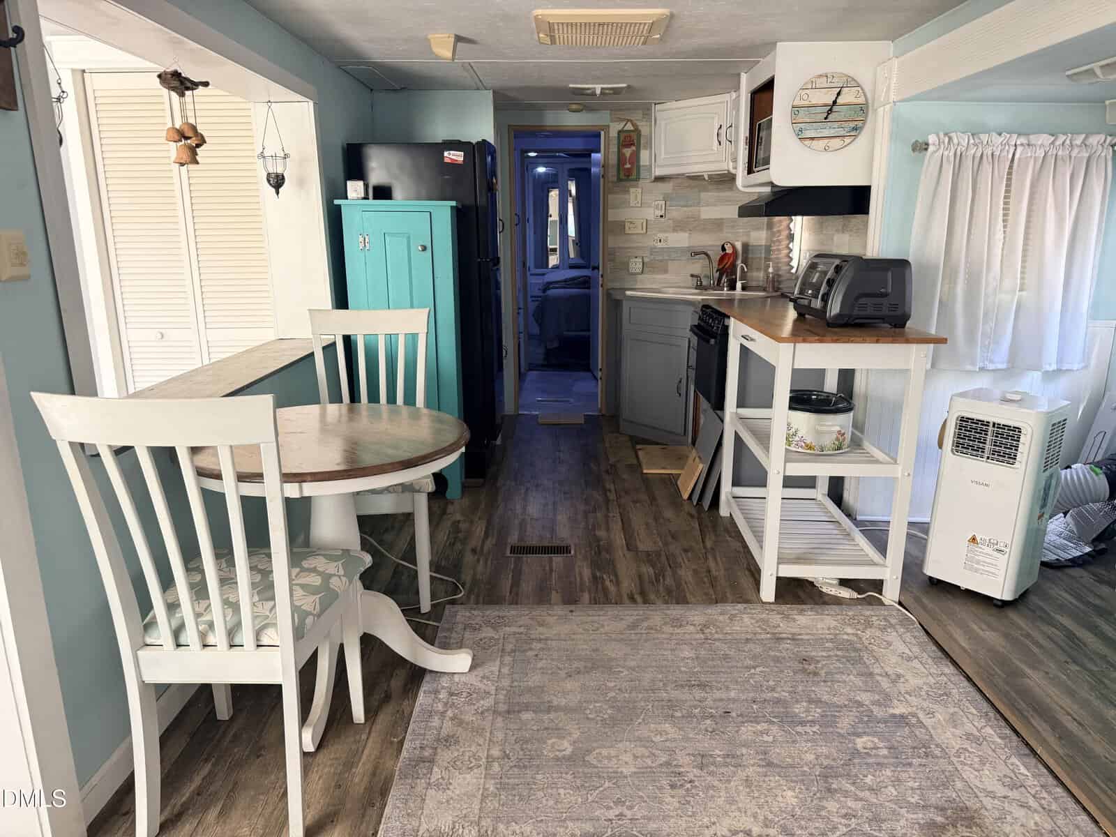 A cozy, light-filled kitchen and dining area in a small home, featuring a round table with two chairs, turquoise cabinet, compact kitchen appliances, and wood-look floors leading to a hallway and bedroom.