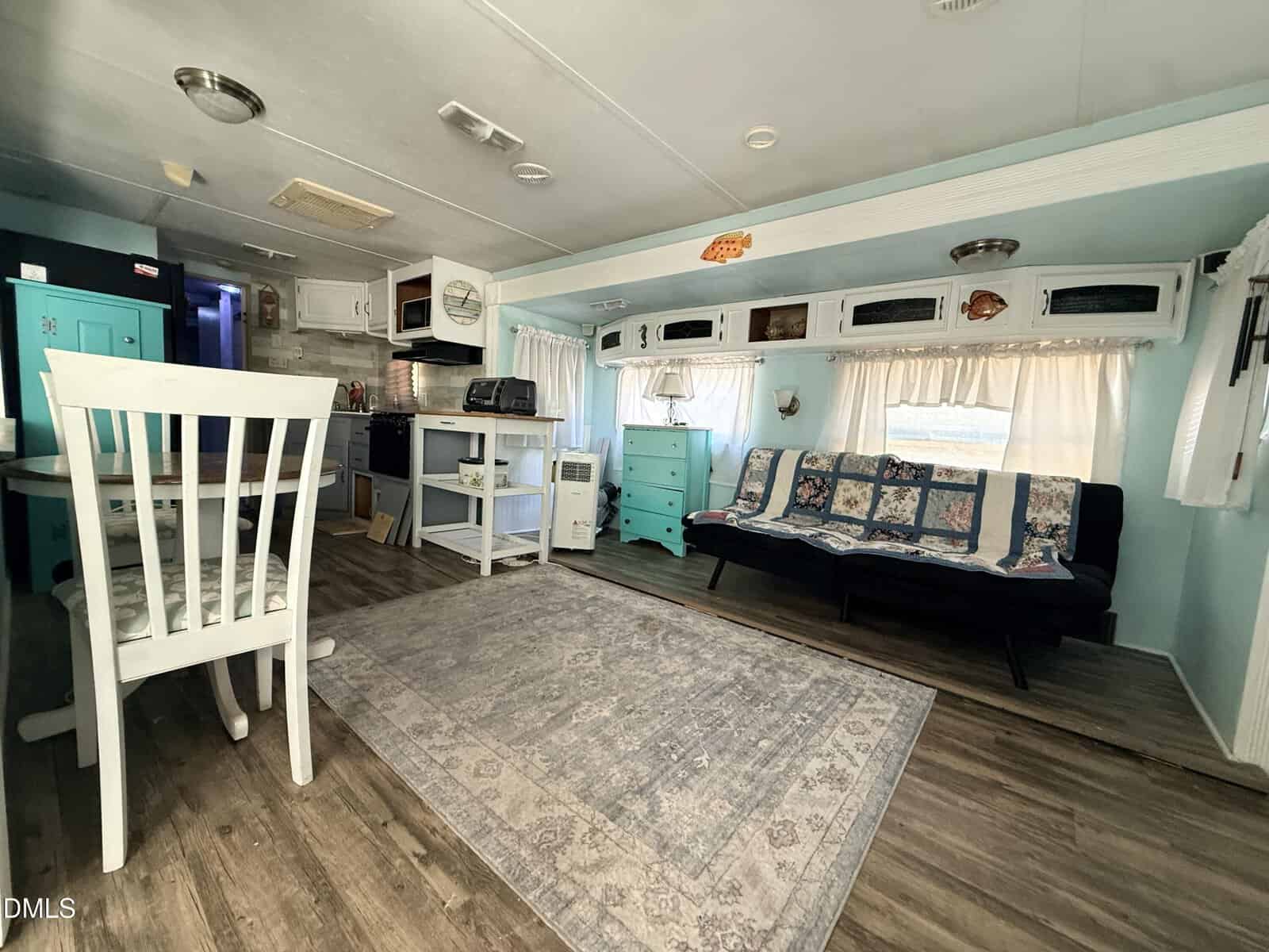 Cozy RV interior with wood flooring, a dining table and chairs, a futon sofa with a quilt, turquoise drawers, kitchen area, white cabinets, and large windows with curtains. The space has a light, airy feel.