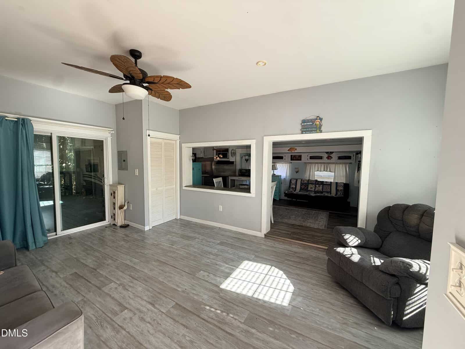 A bright living room with light gray walls, wood-look tile floors, a ceiling fan with leaf-shaped blades, a recliner chair, sliding glass doors with teal curtains, and two large interior windows.