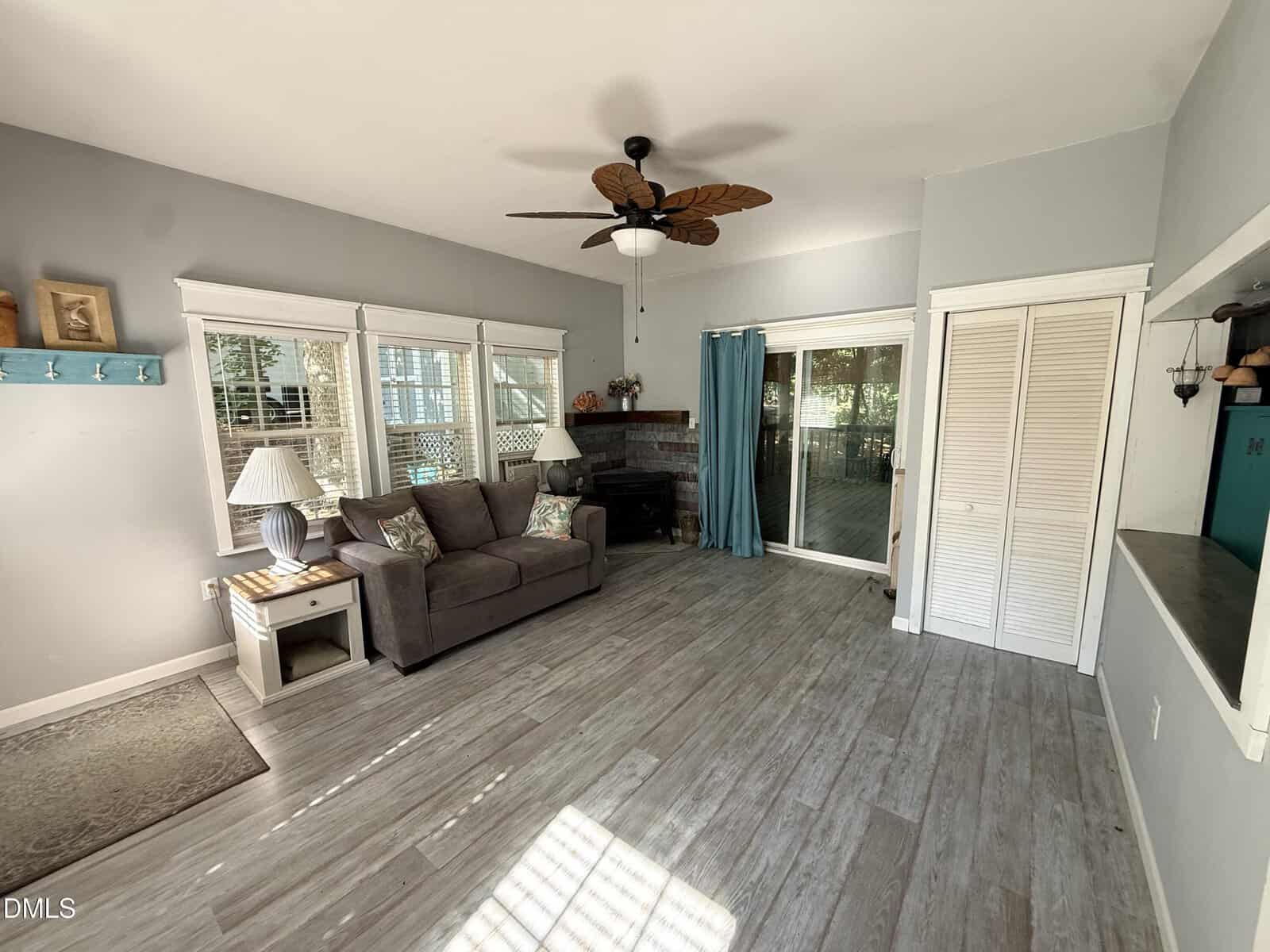A bright living room with gray wood floors, a gray sofa, white side tables with lamps, a ceiling fan, large windows, sliding glass doors with teal curtains, and a white closet. Light streams through the windows.