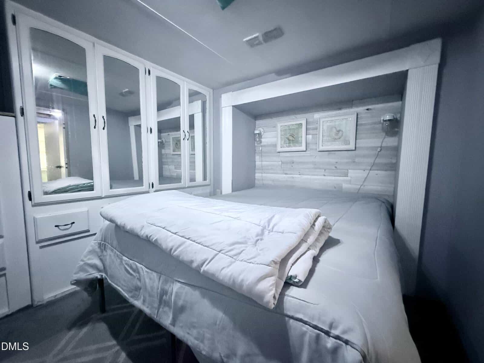A modern bedroom with a large bed, white bedding, built-in mirrored closet, gray walls, three framed artworks above the headboard, and small wall lamps on each side.