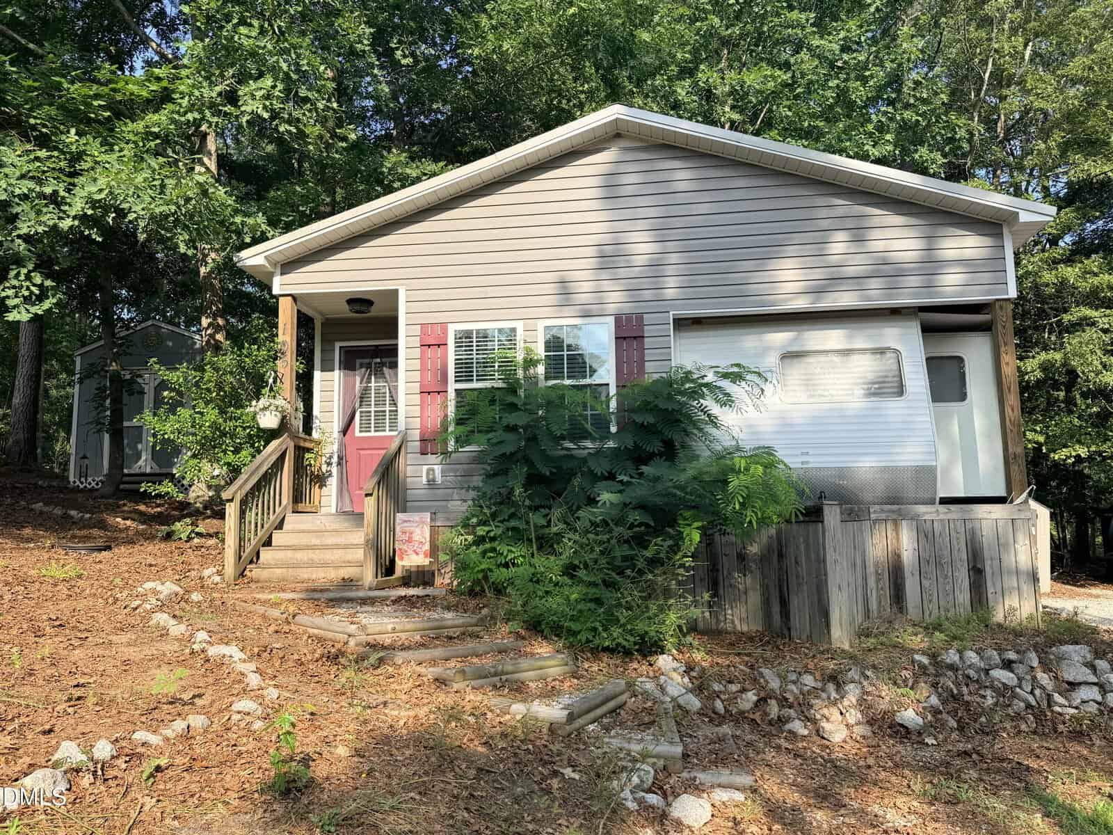 Small beige house with red shutters, front porch, and steps leading up. Overgrown bushes and trees surround the home at 105 Cimarron Drive in Louisburg. A gravel path and patchy lawn are visible in the foreground.