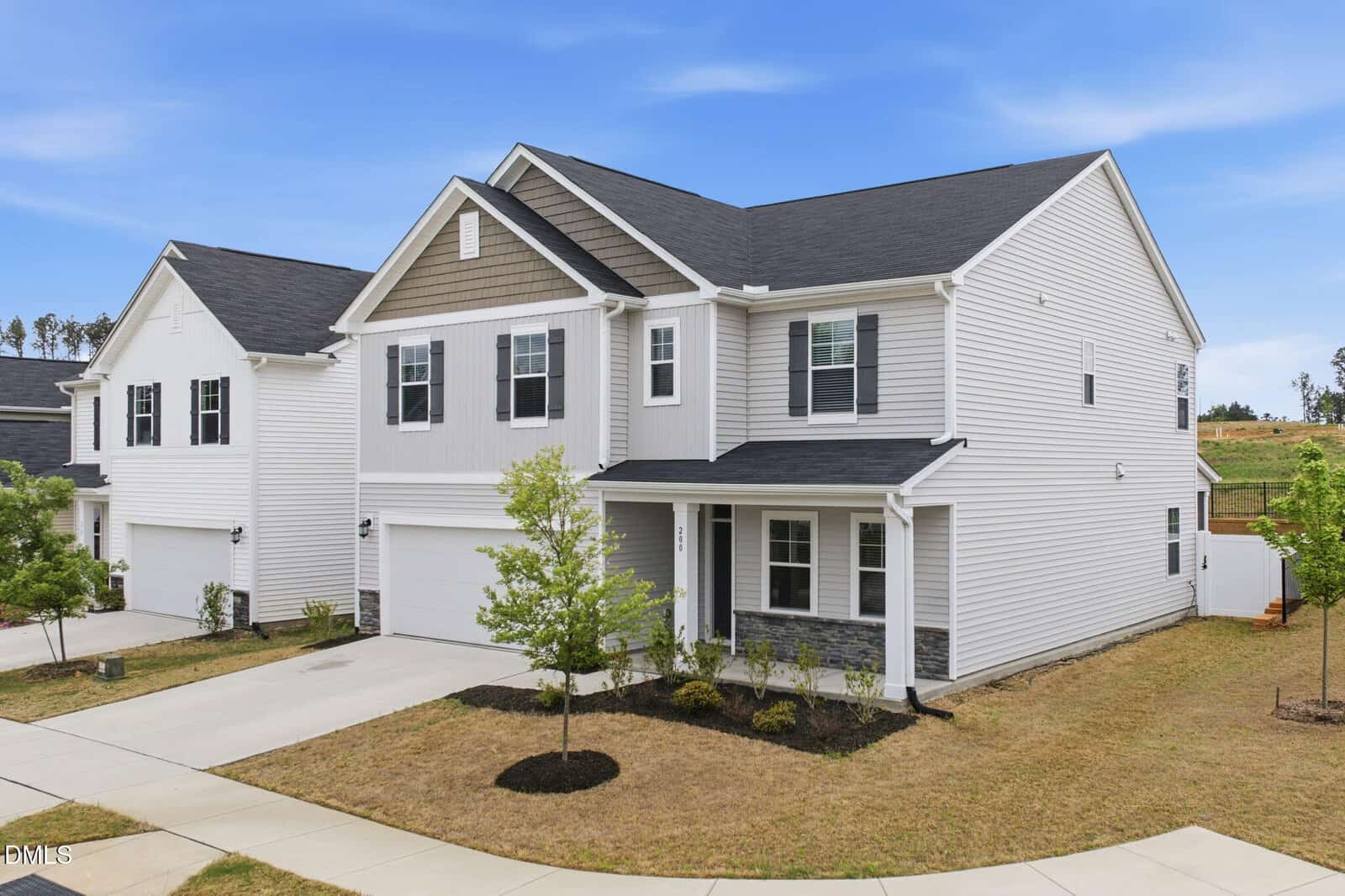 A modern two-story white house with black roof, double garage, small front porch, and young trees planted in the front yard. The lawn is dry, and the sky is clear and blue.