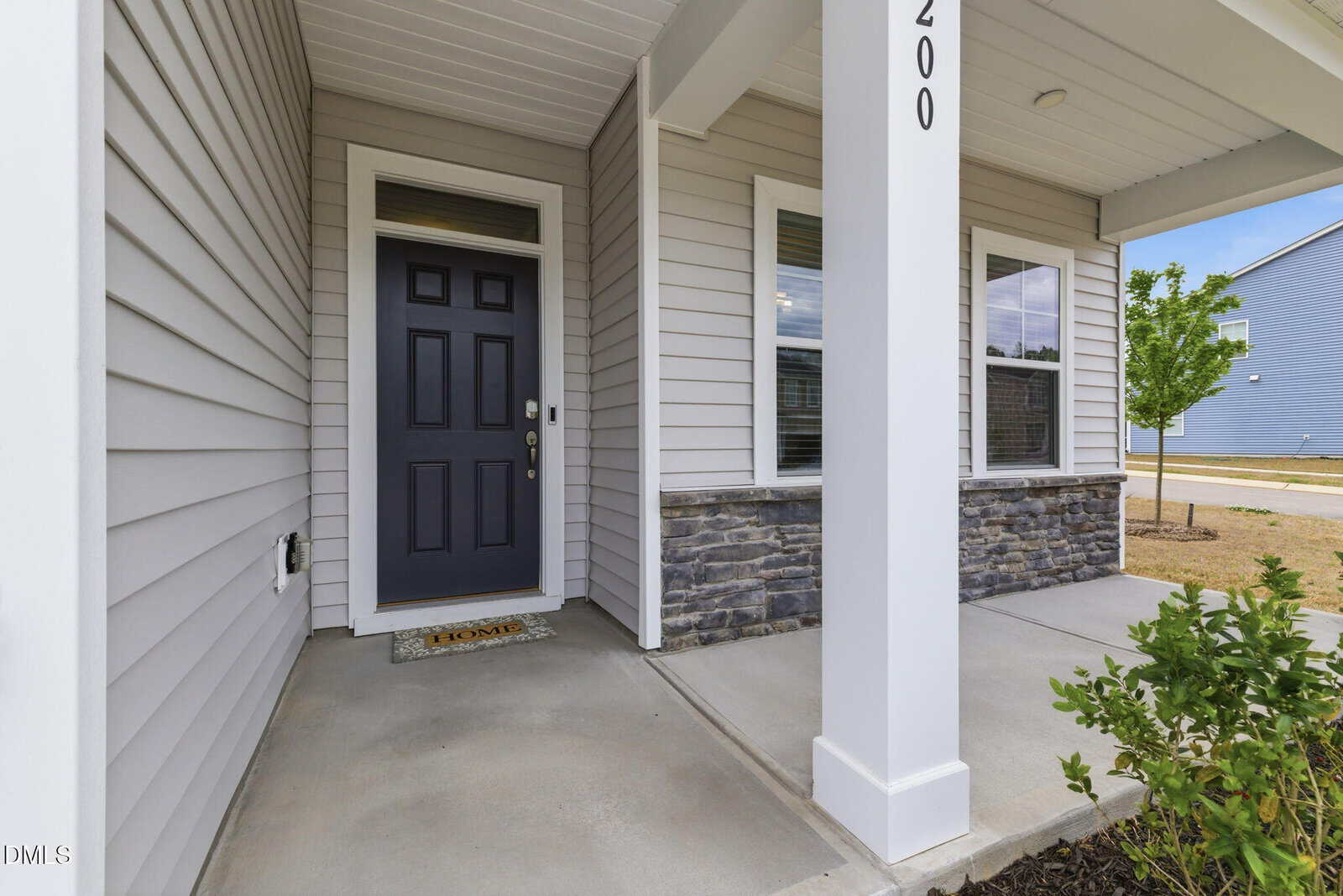 A covered front porch of a modern house with gray siding, a dark front door, stone accents, a welcome mat, and a small landscaped area with young plants. House number "200" is visible on a white column.