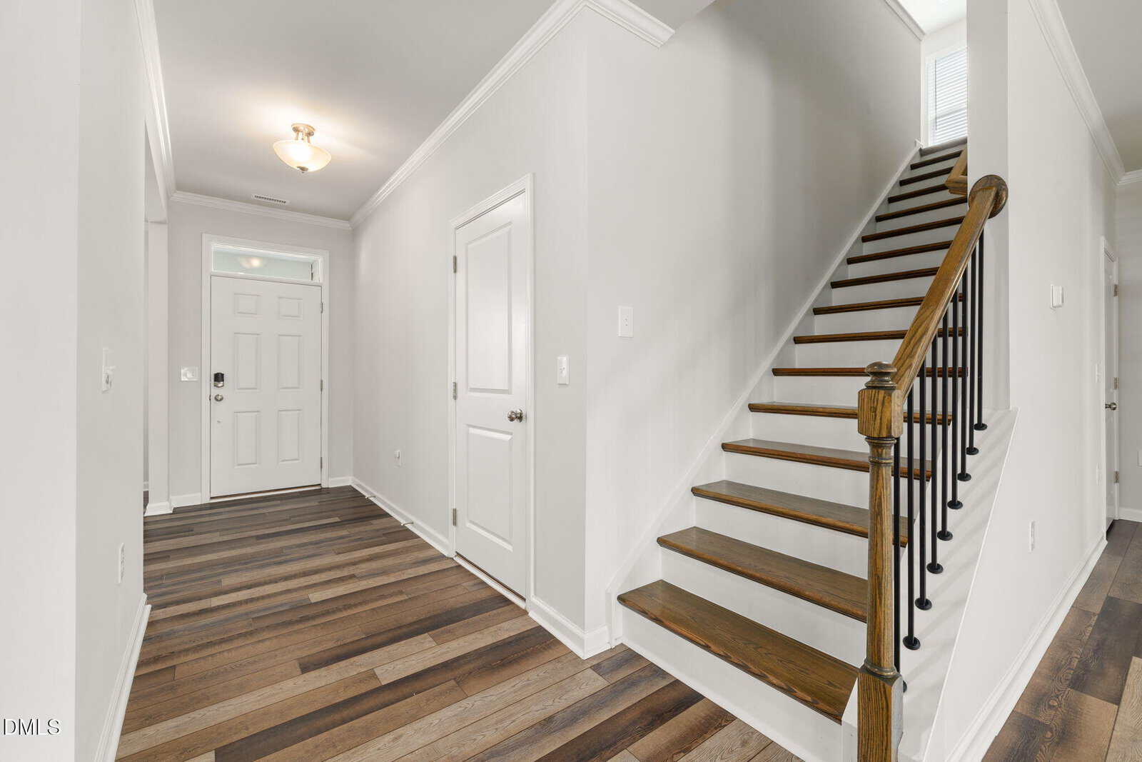 Bright hallway with wood flooring, white walls, and crown molding. There is a staircase with wooden steps and black spindles on the right, and a white front door with an adjacent closet on the left.