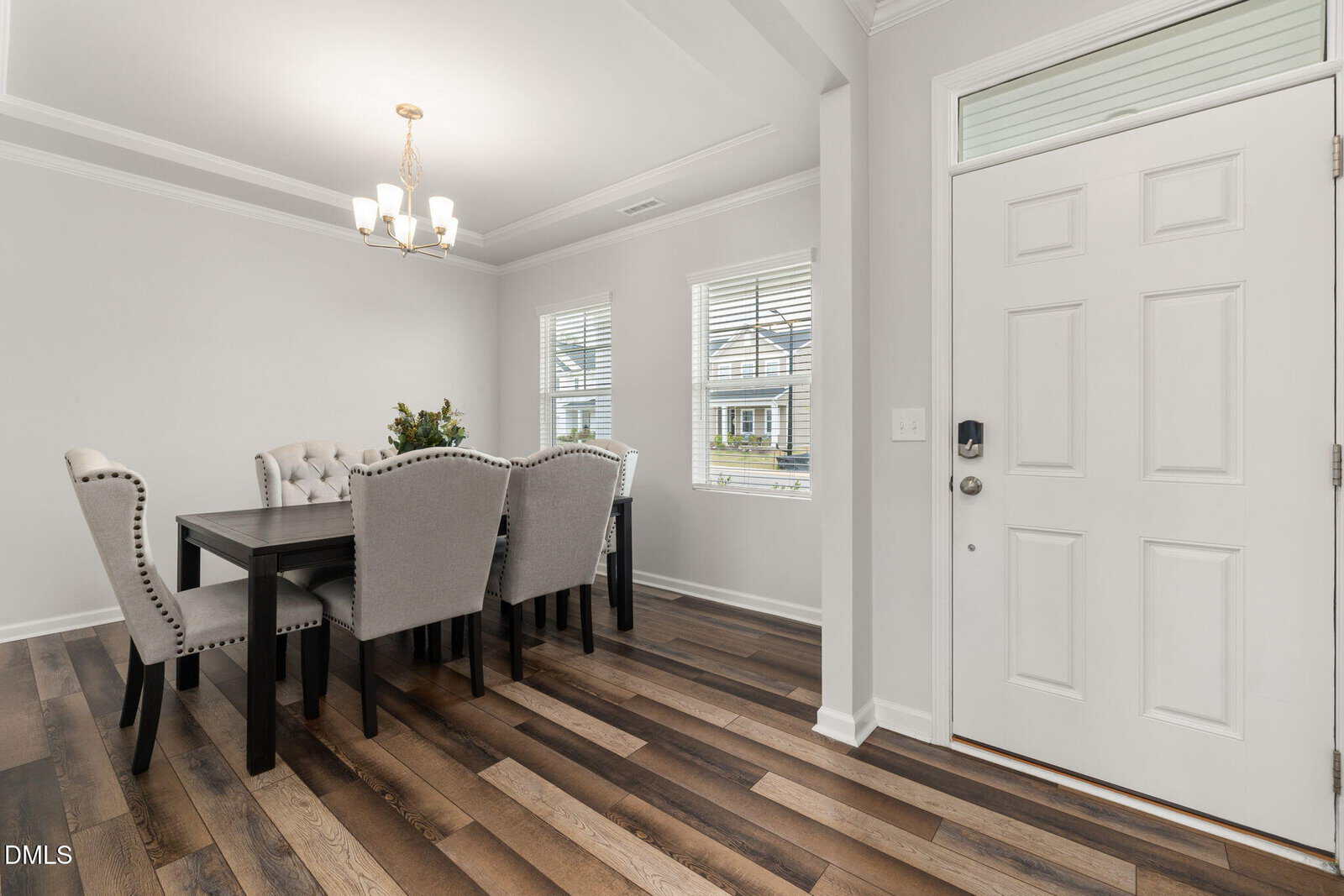 A bright, modern dining room with a dark wooden table, six light upholstered chairs, wood flooring, white walls, two windows, and a white front door with a transom window above it.