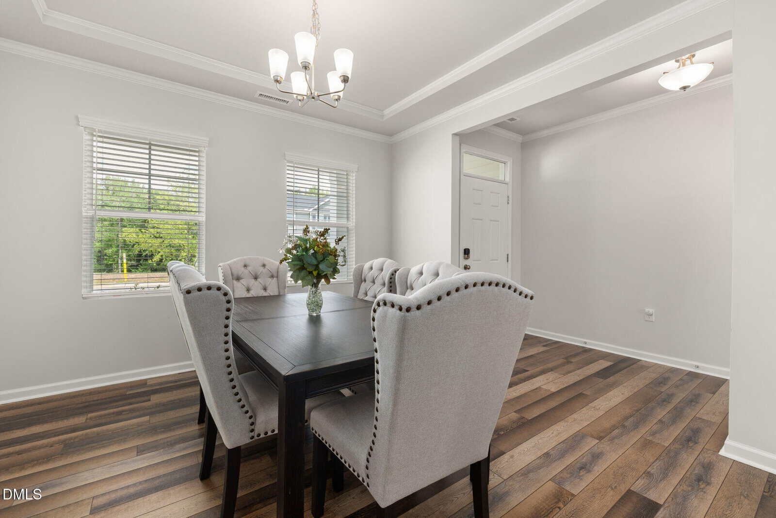 A bright dining room with a black table, six tufted gray chairs, a flower vase centerpiece, wood-style flooring, and two windows with blinds. The entryway with a white door is visible in the background.