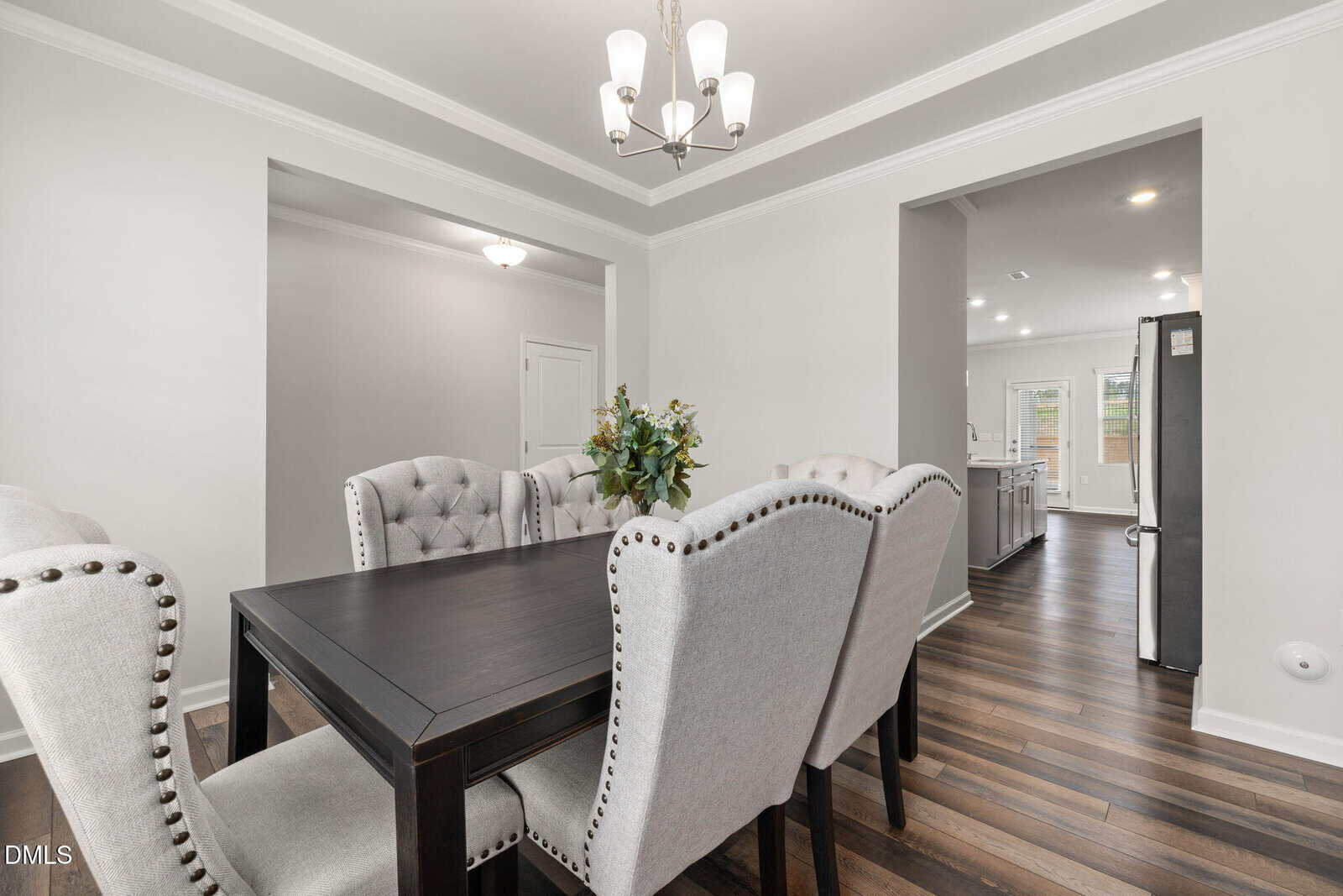 A modern dining room with a dark wooden table, six tufted gray chairs, a floral centerpiece, and a chandelier overhead. The space opens to a kitchen with wood flooring and white walls.