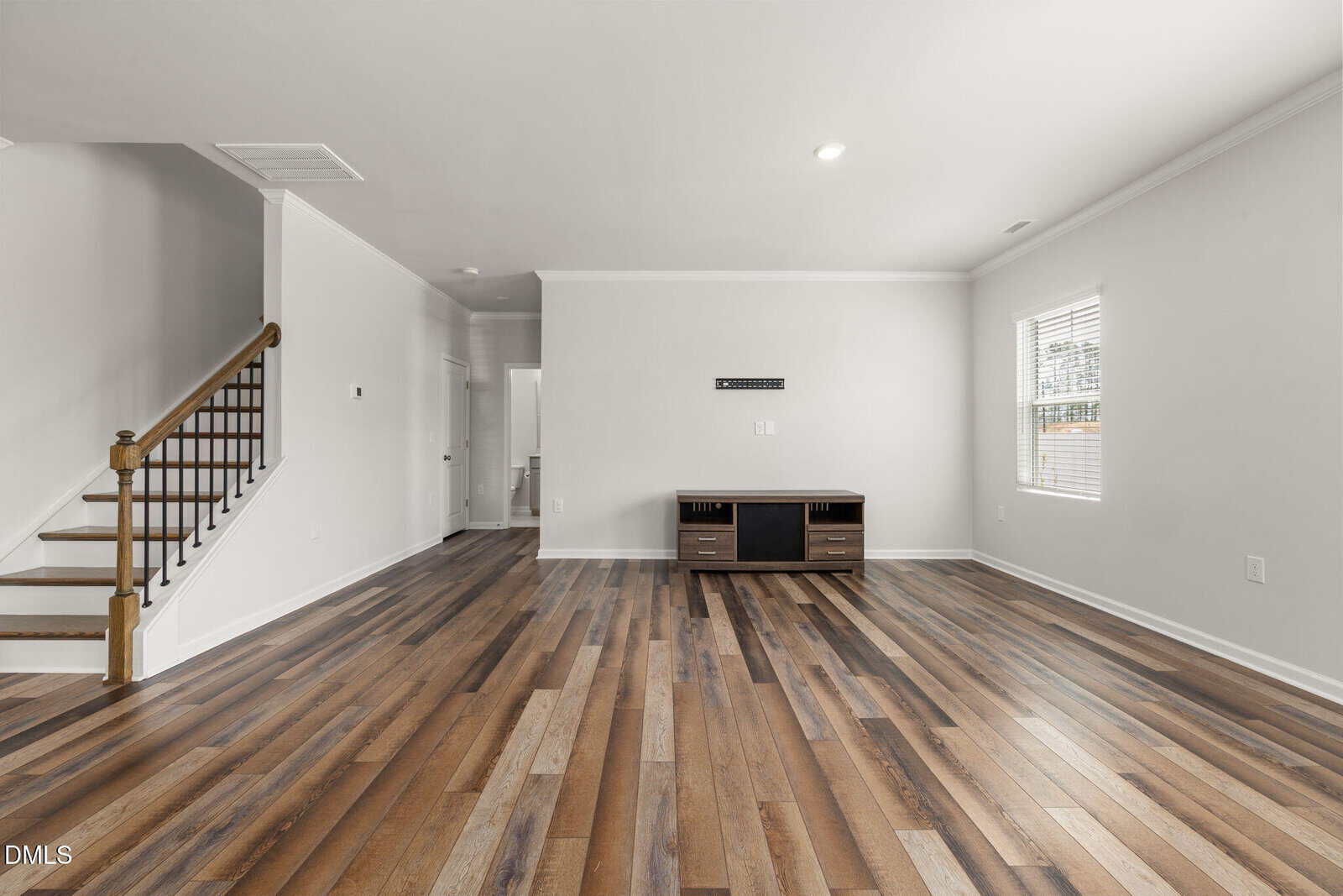 Spacious living room with wood flooring, white walls, a window, staircase, and a TV stand against the far wall. Room is empty and well-lit with natural and ceiling light.