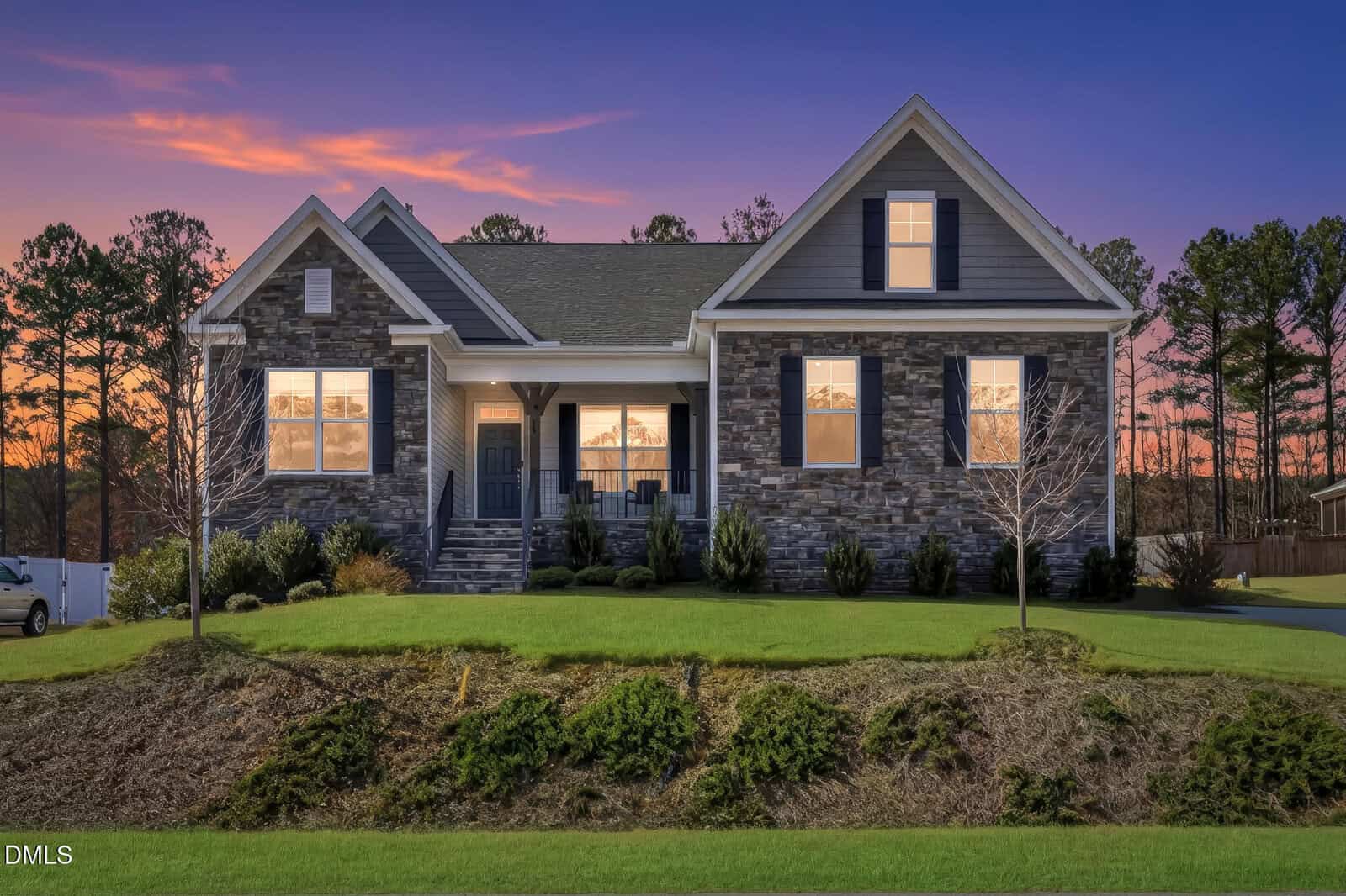 A modern single-story house at 30 Ironwood Boulevard in Youngsville features a dark stone and siding exterior, large windows glowing with warm light, and sits on a grassy lawn at sunset surrounded by trees.