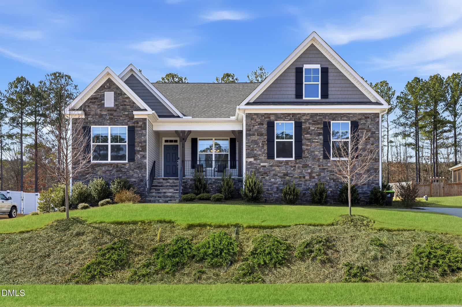 A modern single-story house with a brick and grey siding exterior, large windows, a front porch with columns, surrounded by green grass and landscaped shrubs, against a backdrop of tall trees and a blue sky.