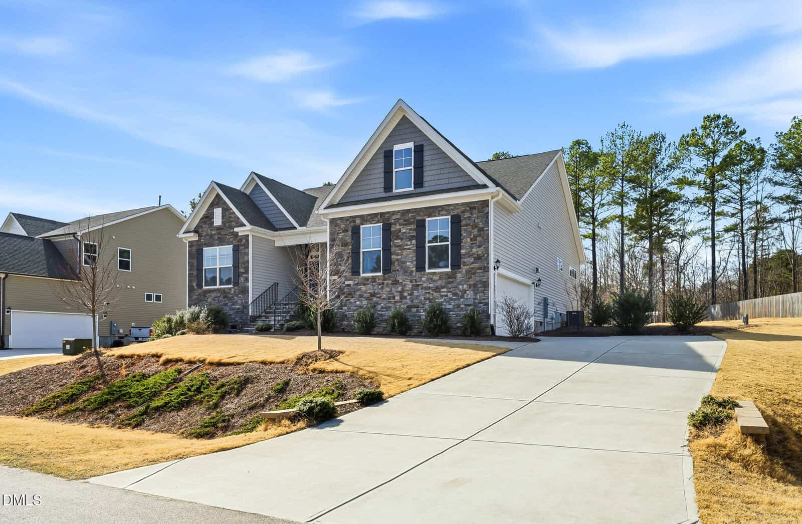 A modern two-story house with stone and siding exterior, a large driveway, landscaped front yard with trimmed grass, small trees, and shrubs, set in a suburban neighborhood with trees in the background.