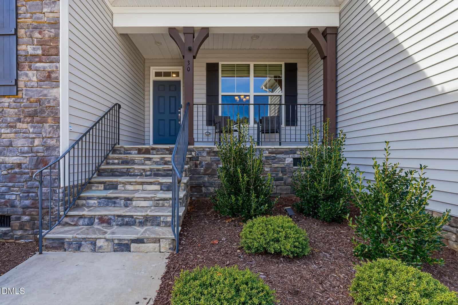 Front view of a house entrance with stone steps, a blue door, white siding, decorative columns, a metal railing, and neatly trimmed green shrubs in a mulched garden bed.