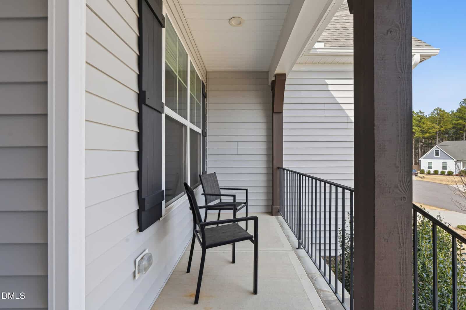 A covered porch with two black chairs, white siding, black shutters, brown posts, and a metal railing overlooking a residential street and houses.