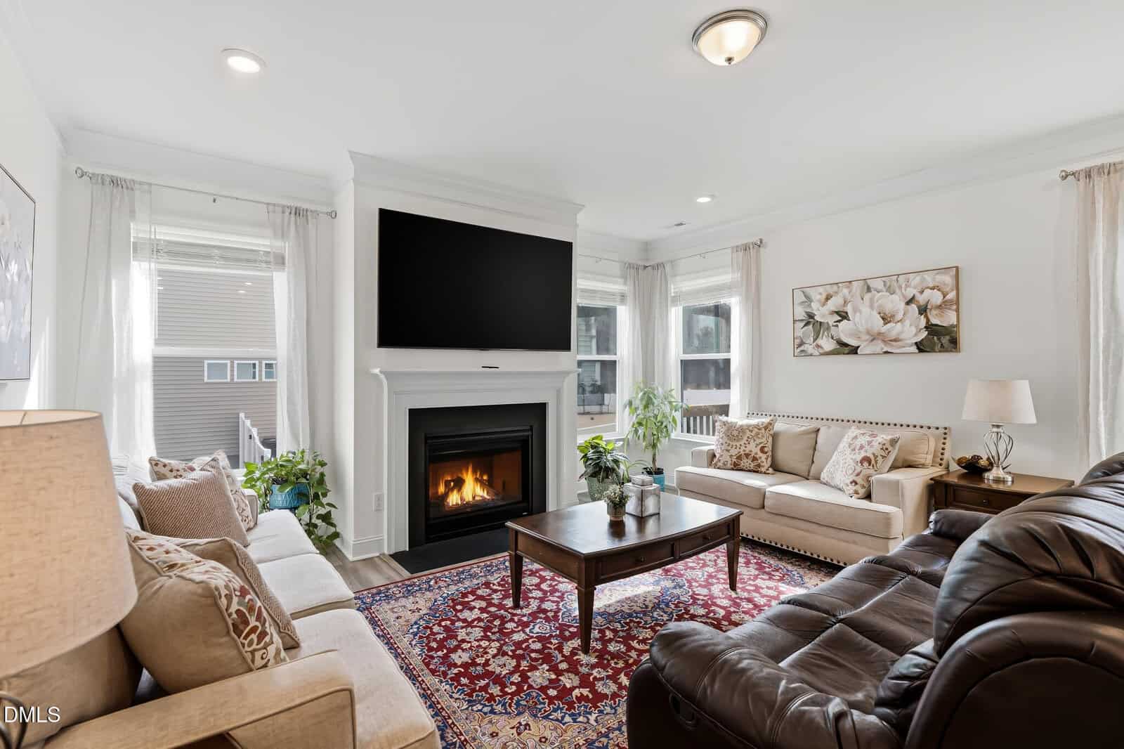 Bright living room with a fireplace, TV above it, beige sofas, a leather recliner, wooden coffee table, red patterned rug, and large windows with white curtains. Plants and artwork decorate the space.