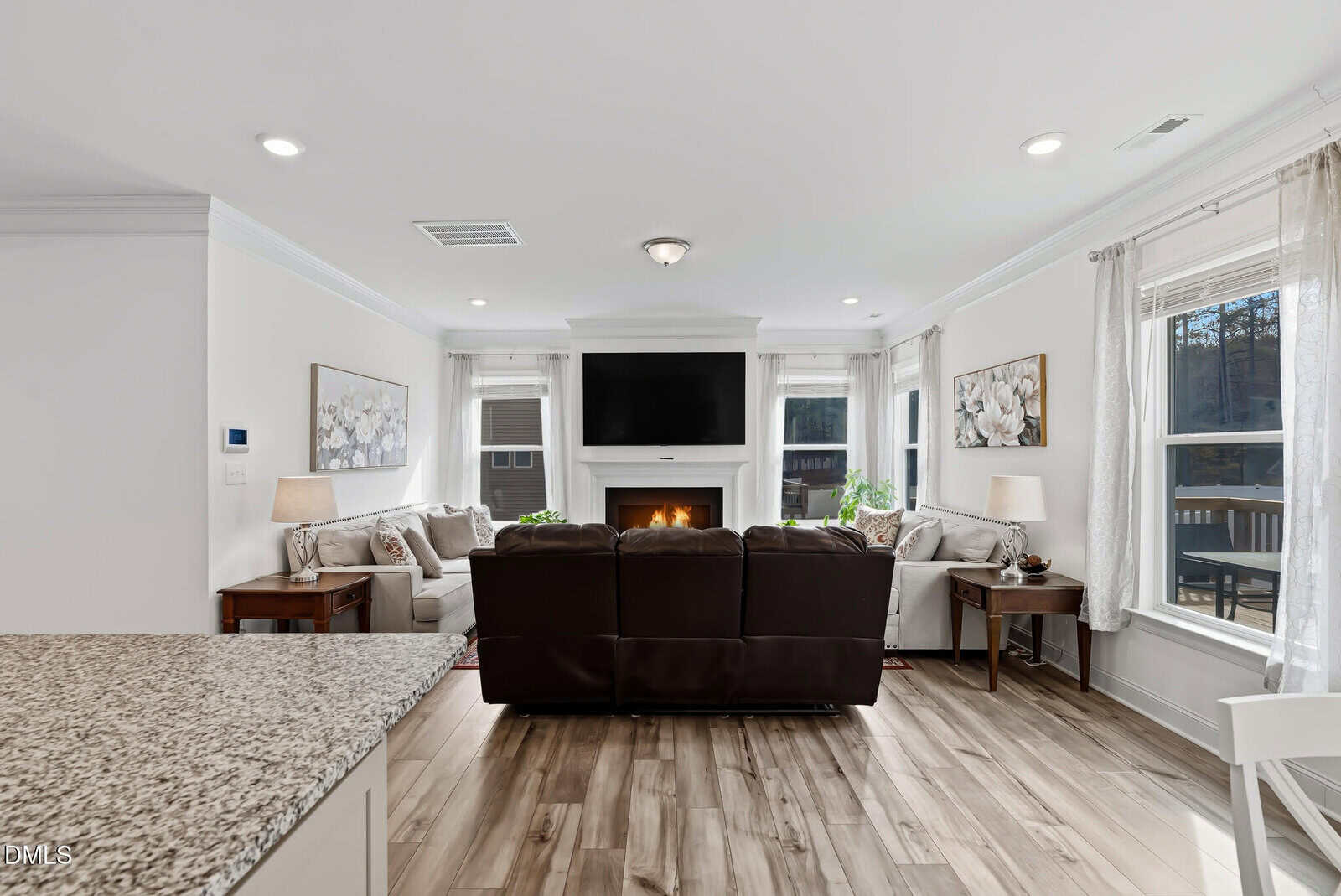 Bright living room with a dark leather sofa facing a fireplace and wall-mounted TV, surrounded by windows with sheer curtains, light walls, floral artwork, and wood flooring.