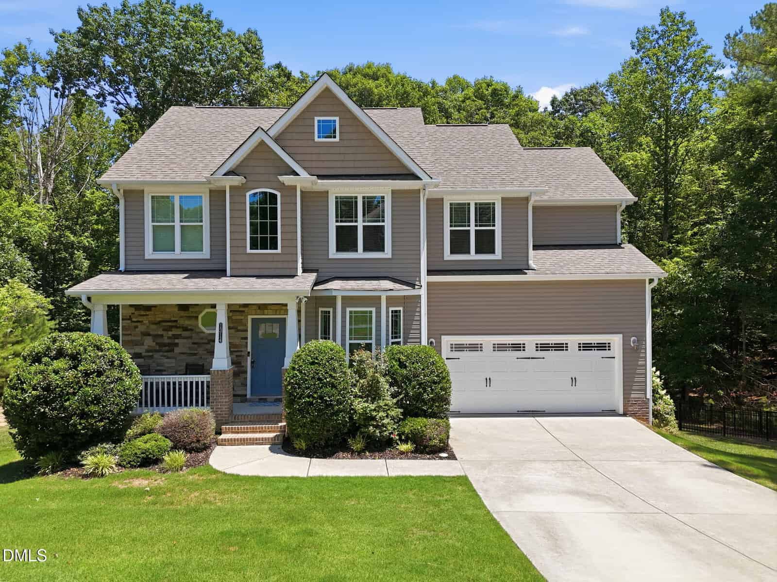 Two-story suburban house at 1314 Sourwood Drive, Wake Forest, with gray siding, stone accents, and a covered front porch. Green shrubs and trees surround the home. A concrete driveway leads to a double garage under a clear blue sky.