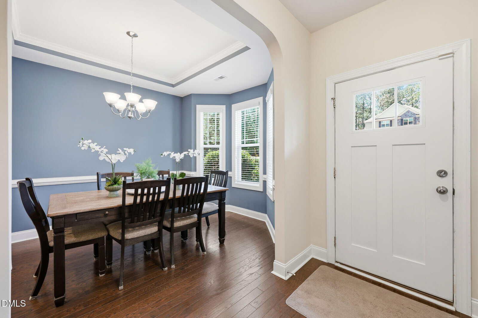 A bright dining room with blue walls, white trim, and dark wood flooring. A wooden table with six chairs is set under a modern chandelier, and three large windows let in natural light near the home's front door.