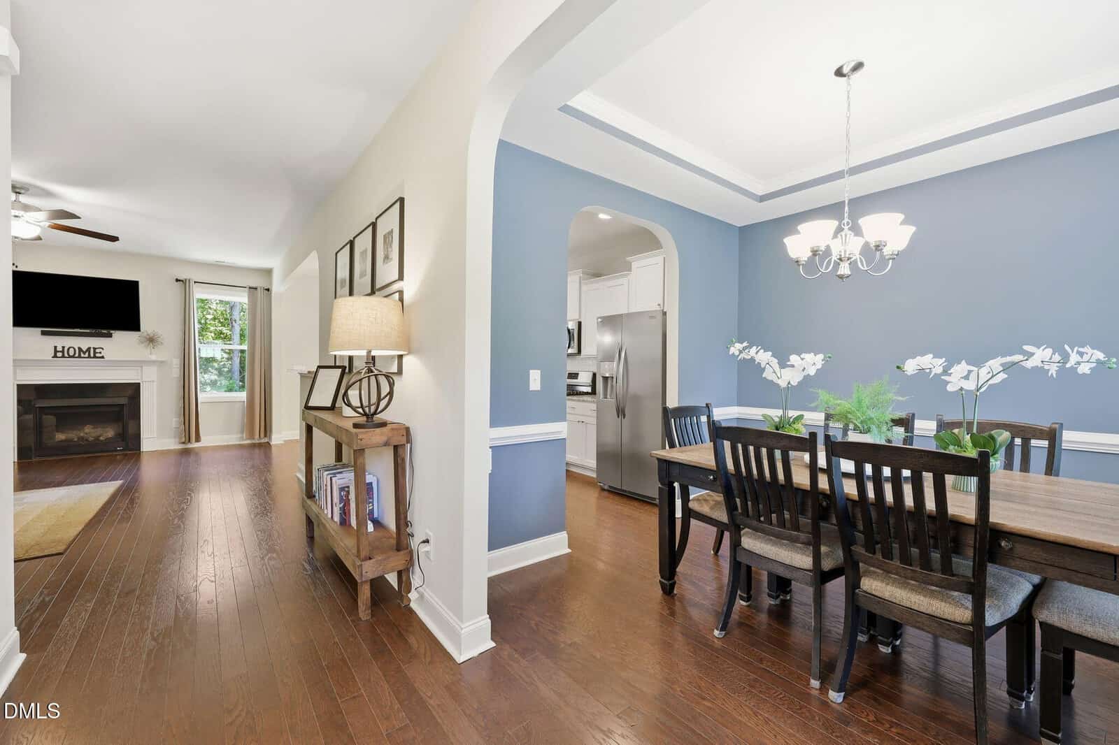 A hallway with wooden floors leads to a living room with a fireplace and a wall-mounted TV. To the right is a dining room with blue walls, a table with six chairs, and a chandelier above. A kitchen is partially visible.