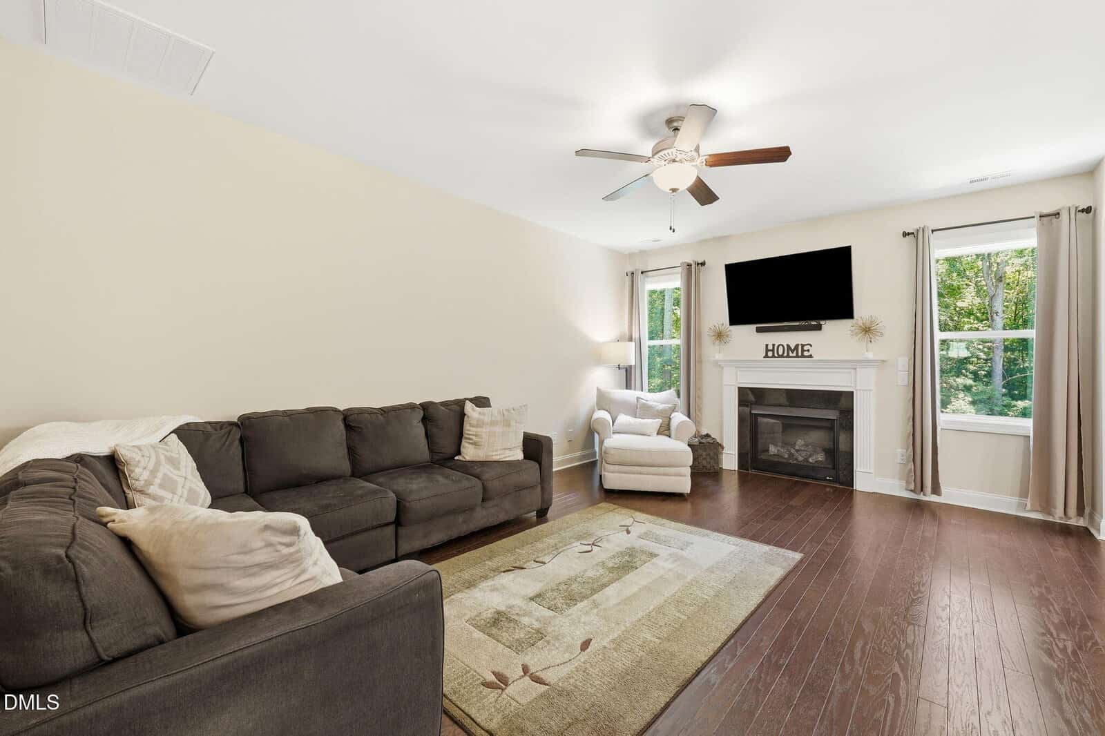 Bright living room with a dark sofa, armchair, fireplace, wall-mounted TV, rug, and large windows with beige curtains. Ceiling fan and throw pillows complete the cozy, inviting space.