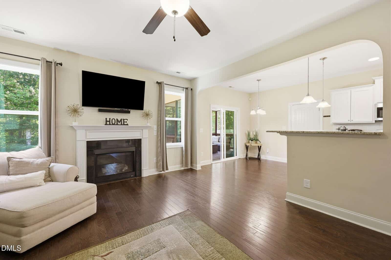 Bright living room with wood floors, a fireplace with “HOME” decor, wall-mounted TV, beige sectional, large windows, and an open view into a modern kitchen with pendant lighting and white cabinets.