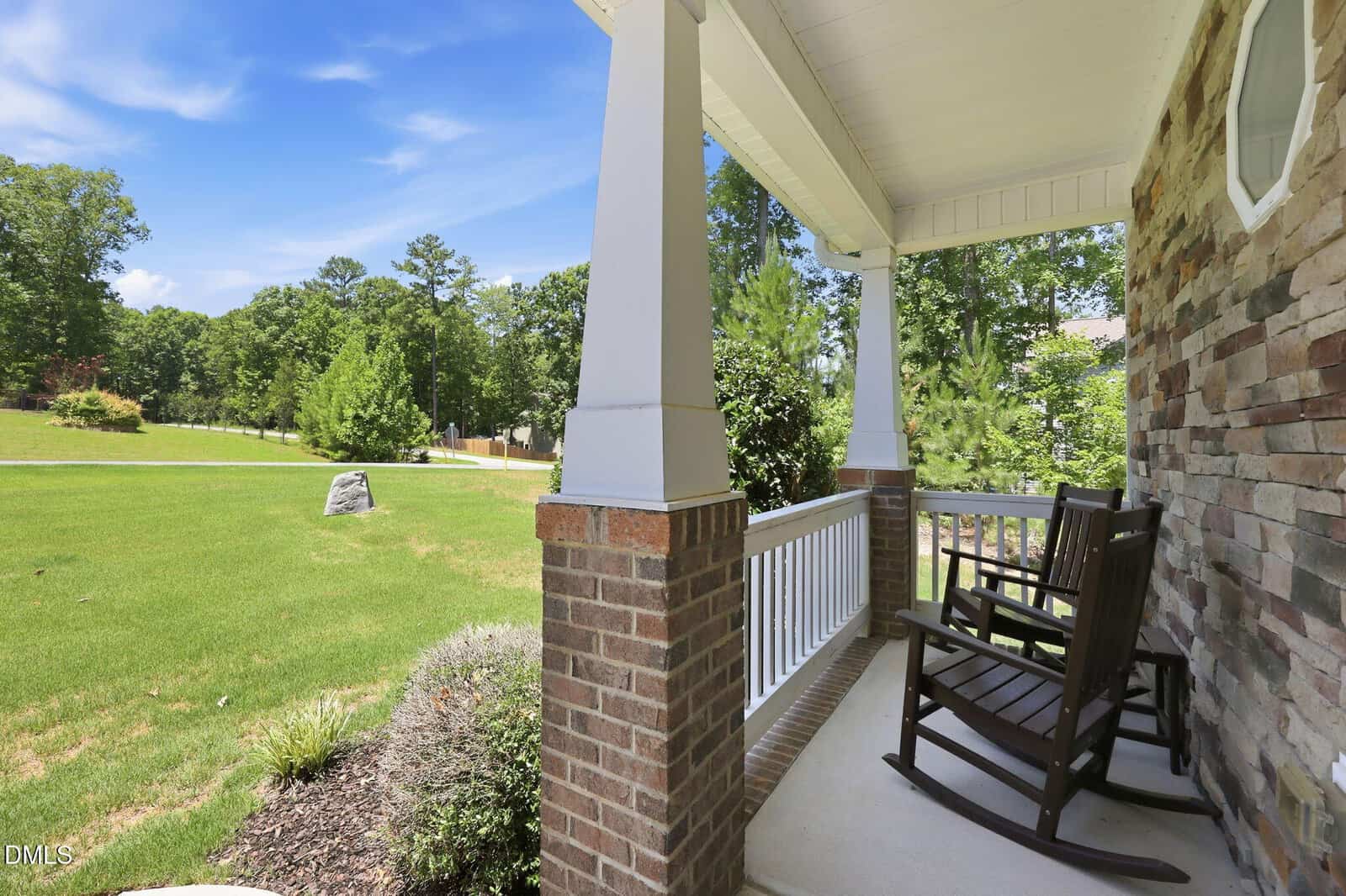 A porch with two wooden rocking chairs overlooks a green lawn and trees under a bright blue sky. The porch has white railings and brick columns, and the house exterior features stonework.