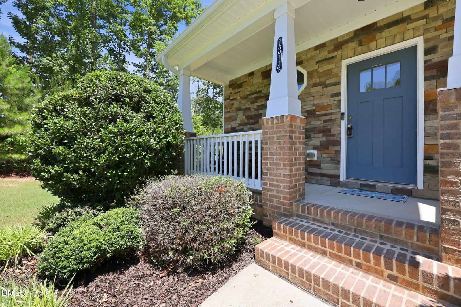 A brick porch with steps leads to a blue front door on a house with stone siding. Bushes and greenery surround the entryway, and a white railing lines the porch. Trees are visible in the background.