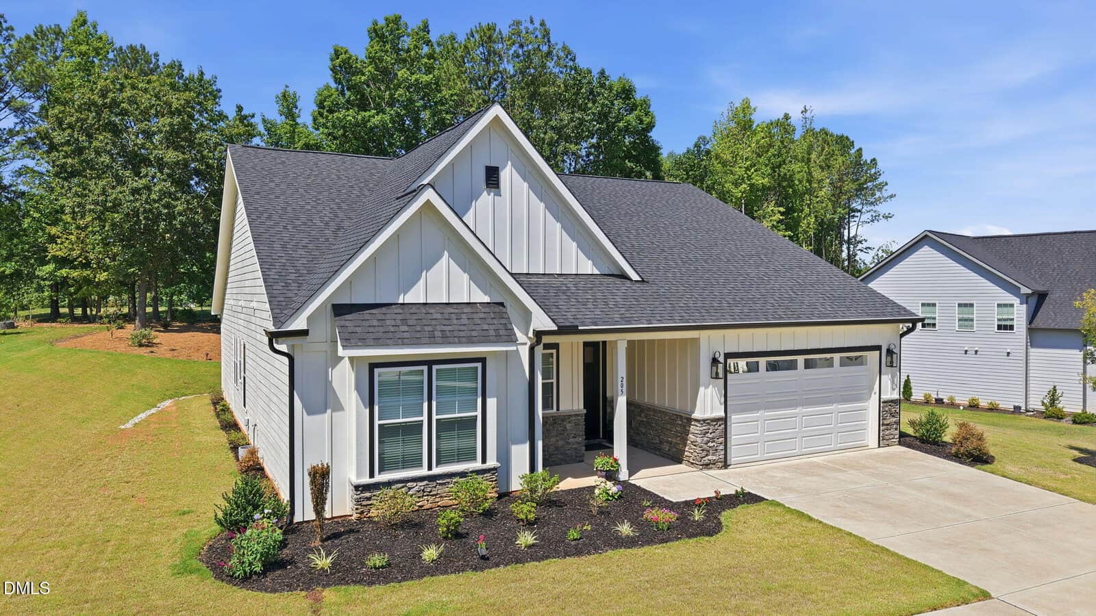 A modern single-story house with white board and batten siding, black roof, stone accents, attached two-car garage, large front windows, manicured lawn, and landscaped garden beds under a clear blue sky.
