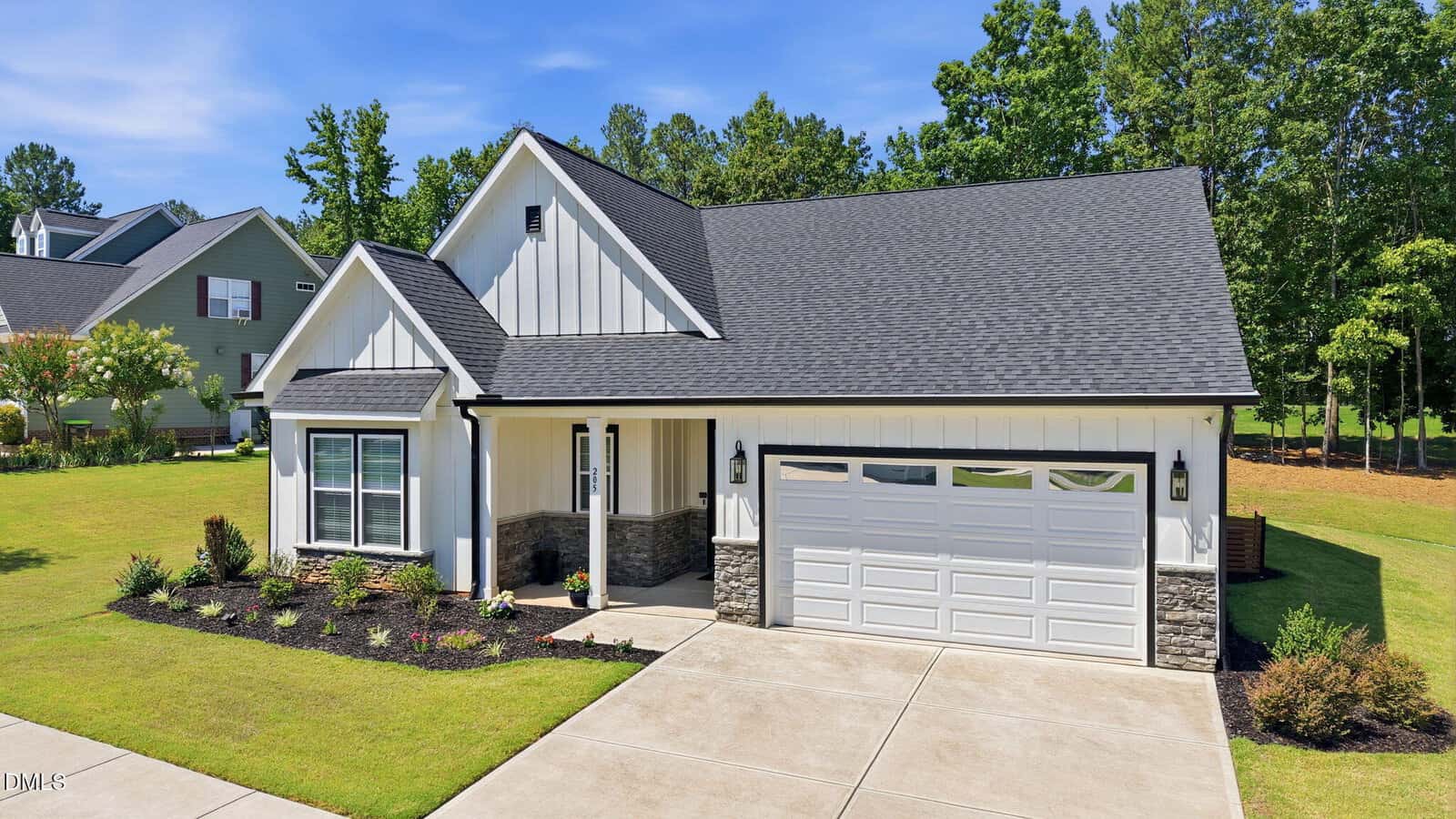 A modern single-story house with white board-and-batten siding, gray stone accents, a dark roof, a two-car garage, and a well-kept lawn and landscaping, set in a suburban neighborhood with trees and clear blue sky.