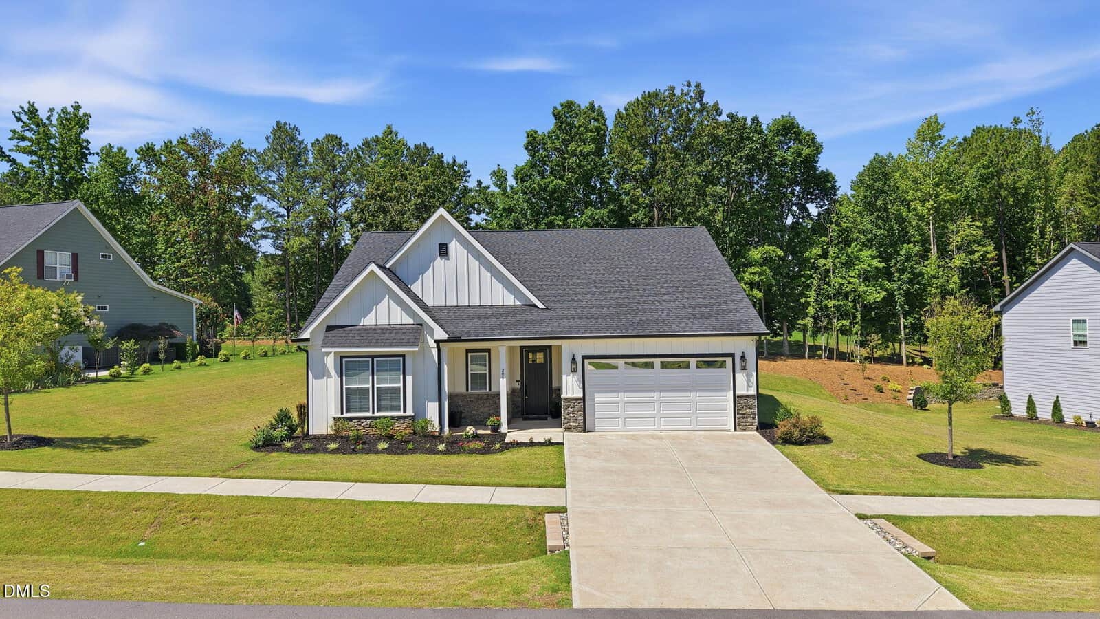 Single-story modern house with white siding, dark roof, and two-car garage. The home has a neat lawn, landscaped garden beds, and a wide driveway, with trees and neighboring houses in the background under a blue sky.