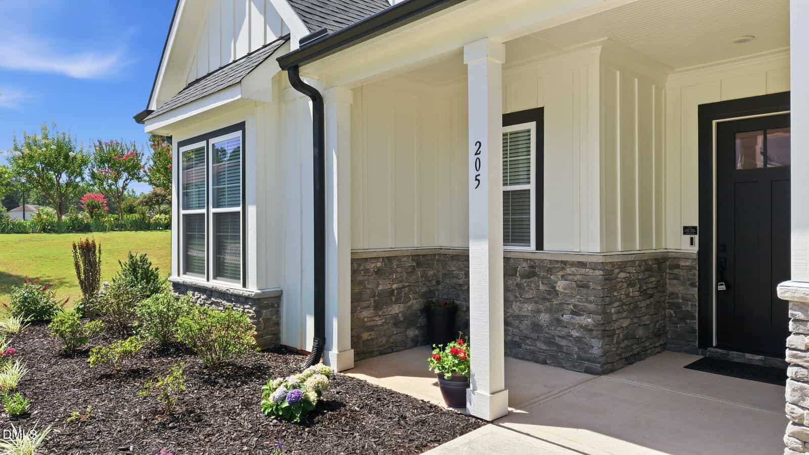 A modern house exterior with white siding, stone accents, and black trim; a covered entryway with the number 205 on a column; landscaped garden with flowers and shrubs in the front yard.